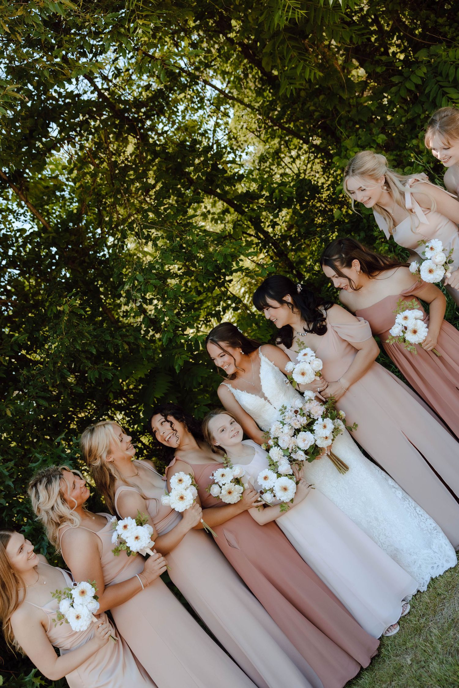 Bride and bridesmaids in blush gowns holding white floral bouquets laughing together outdoors under lush green trees.
