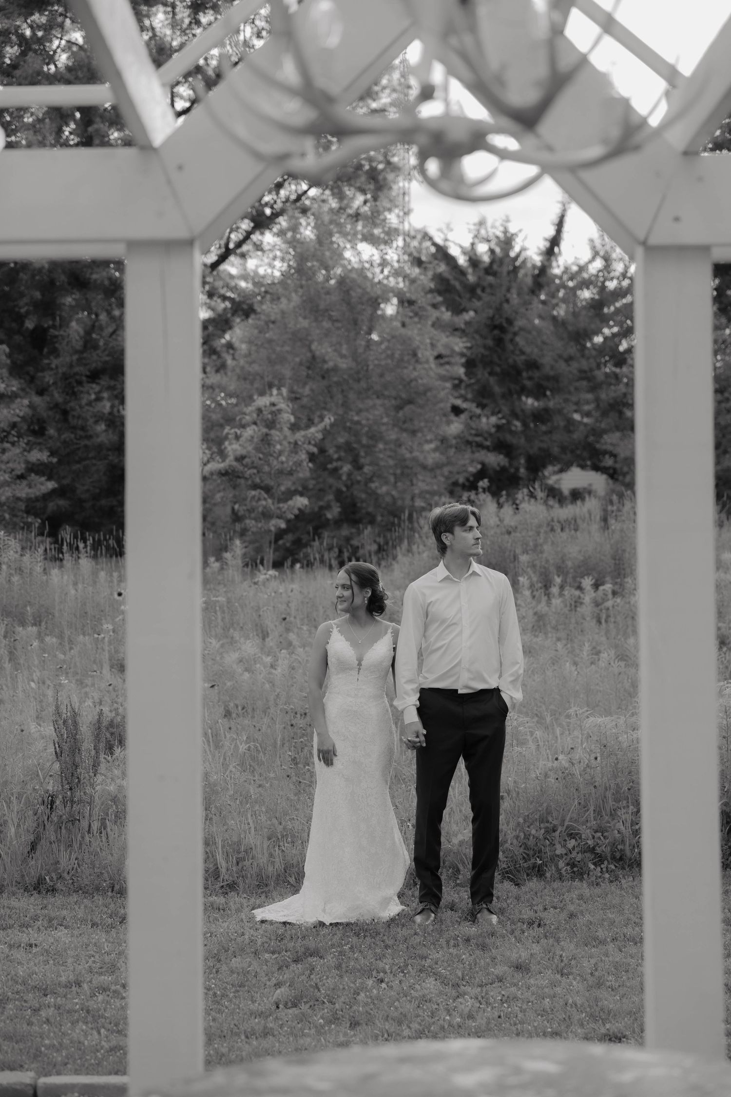 Black and white photo of bride and groom holding hands in a field, framed by a white wooden arbor.