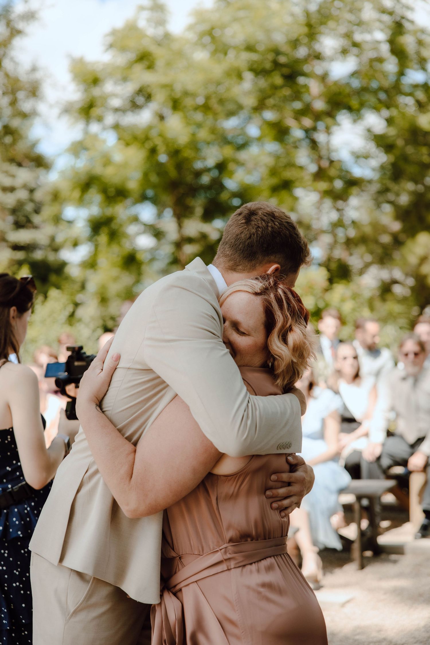 A couple shares an emotional hug during an outdoor wedding ceremony surrounded by guests and lush green trees.