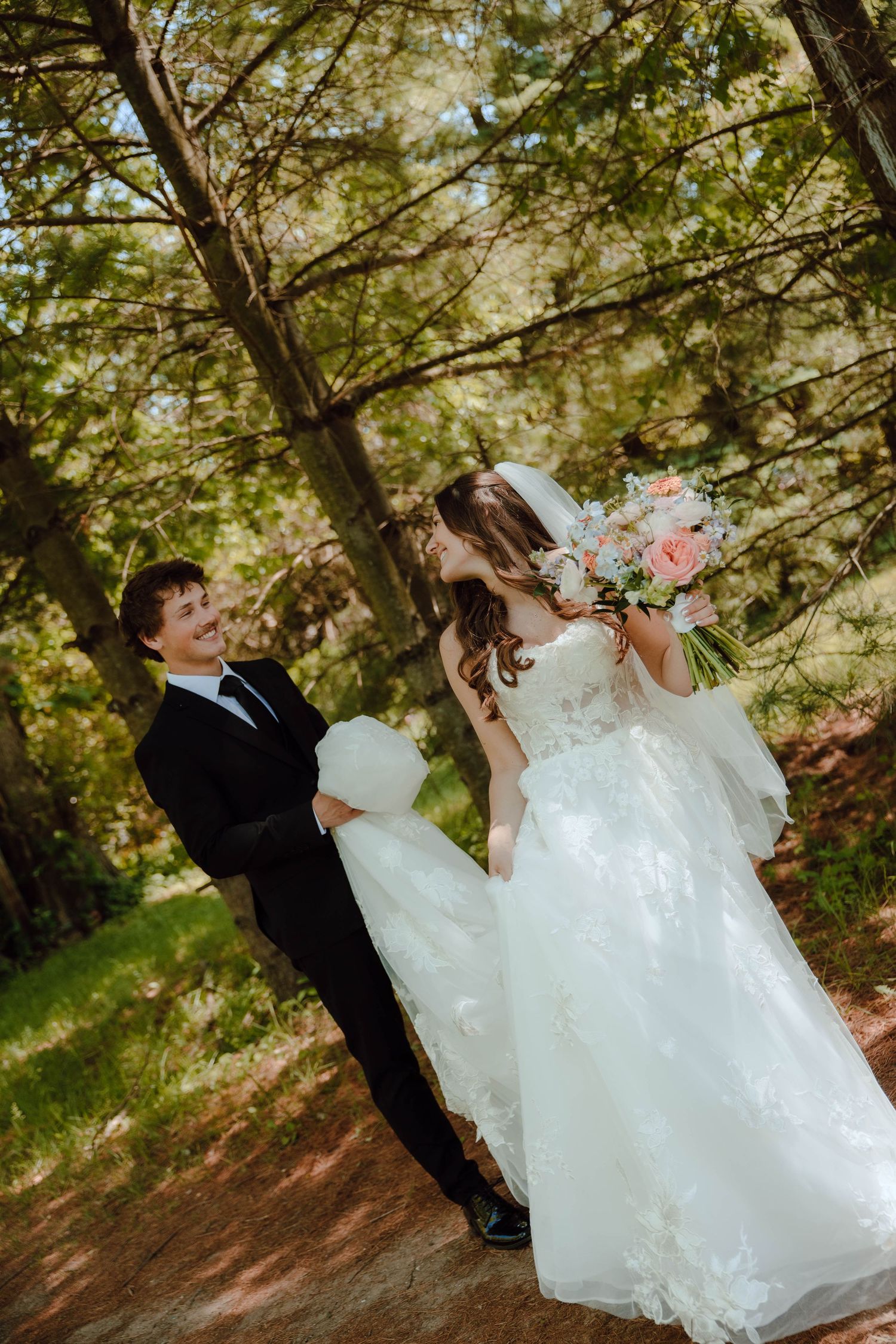 Bride in white lace gown holding floral bouquet laughs with groom in black tuxedo outdoors in sunlit forest setting.