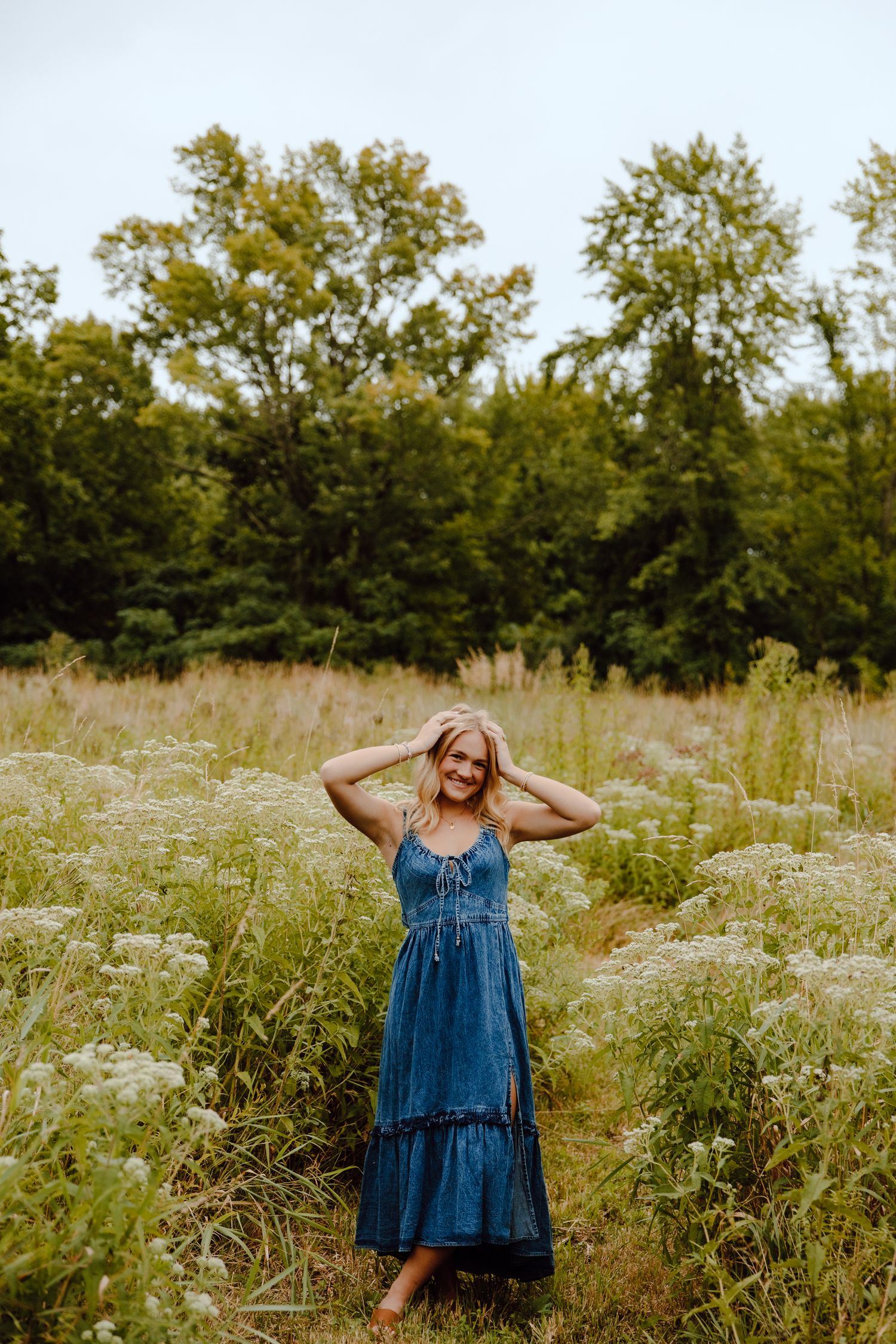 Person in denim dress poses playfully in a wildflower field on a sunny day.