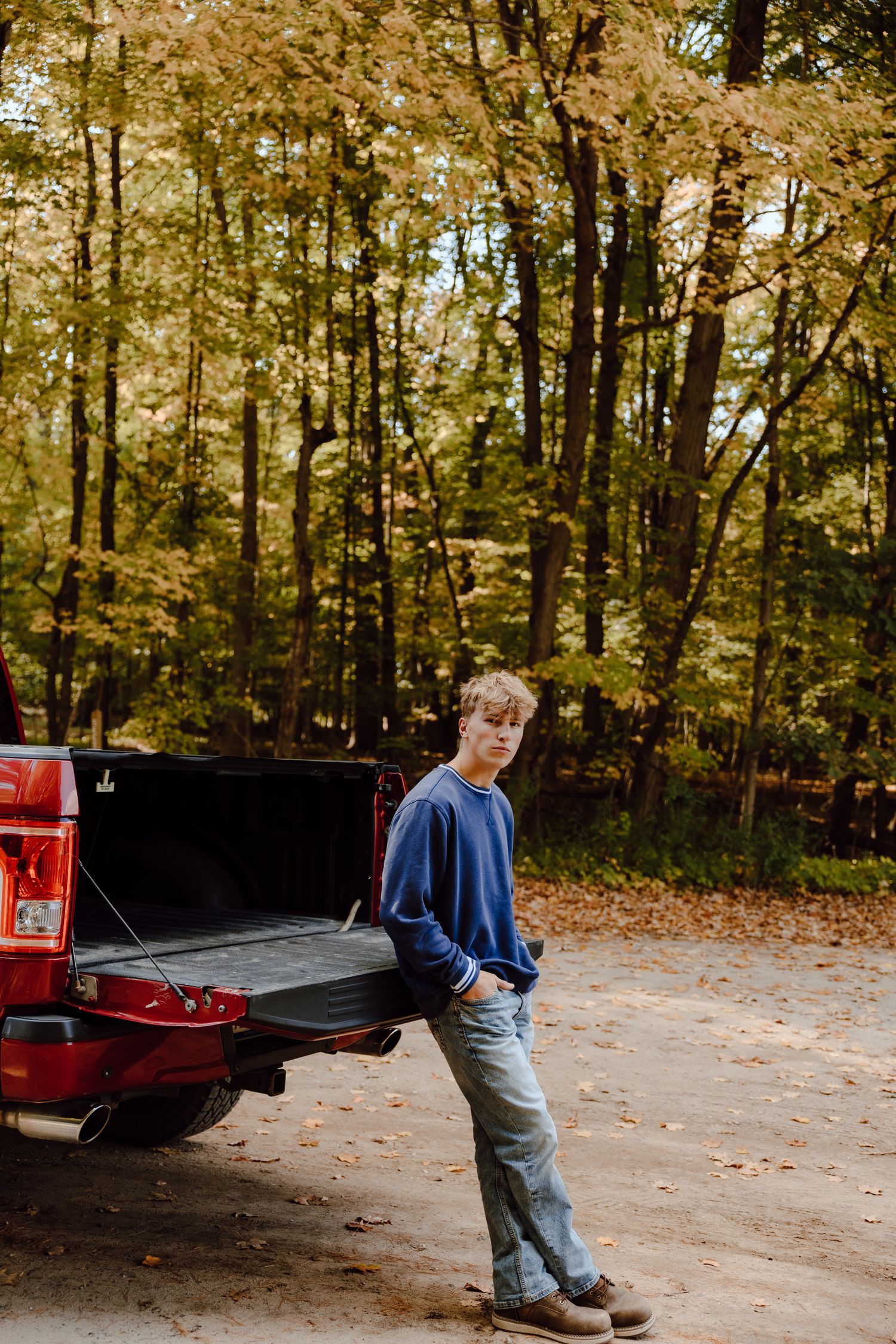 Person in casual attire leaning against a red pickup truck tailgate in a wooded area.