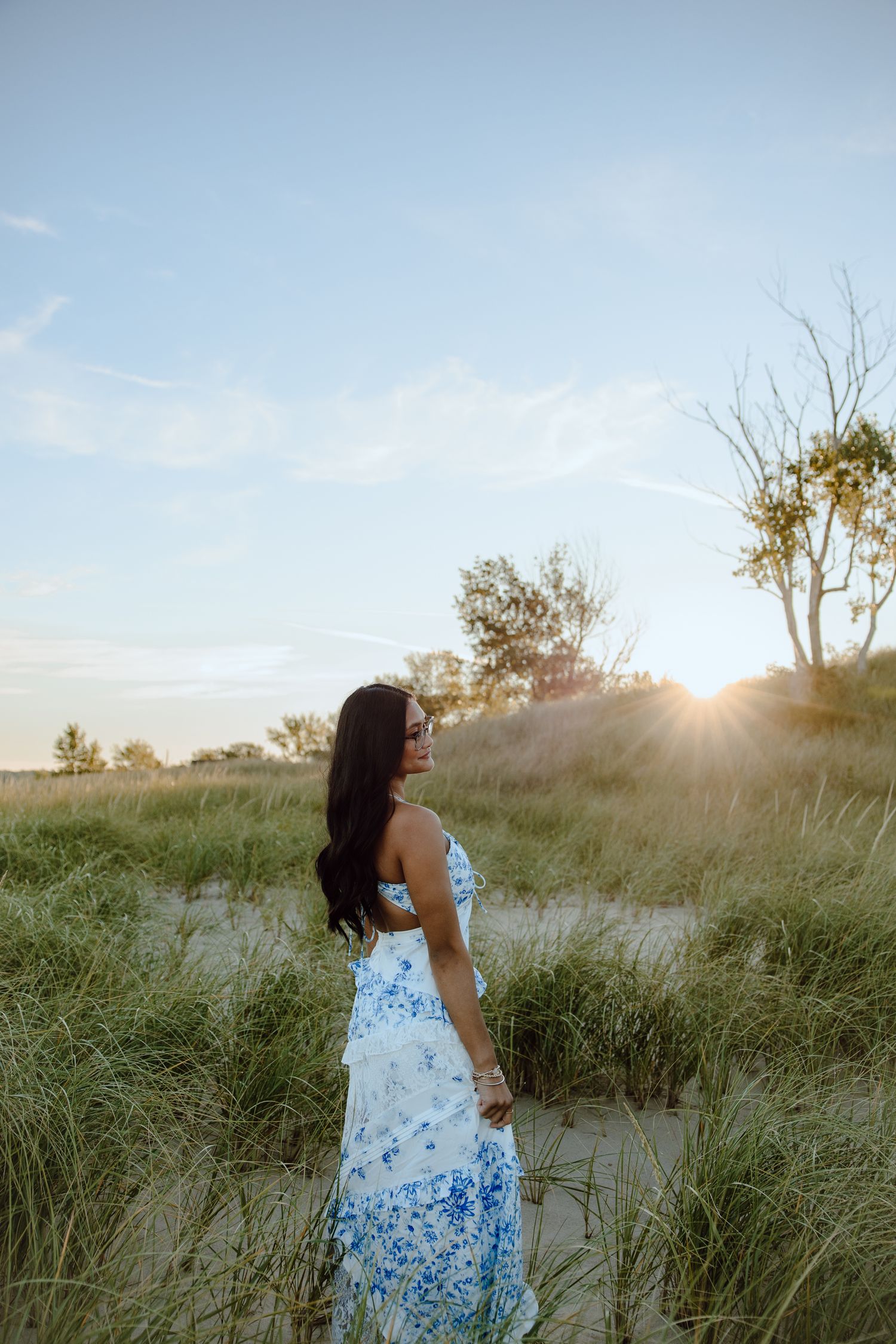 A flowing blue and white dress moves gracefully among coastal dunes at sunset.