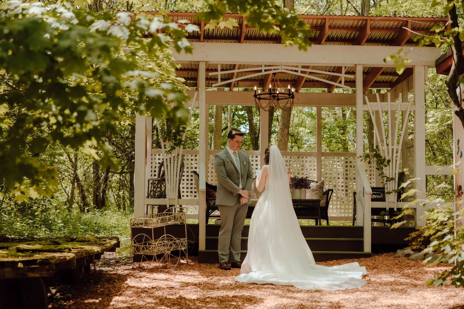 Bride and groom hold hands under a white garden gazebo surrounded by lush green trees at an outdoor wedding ceremony.