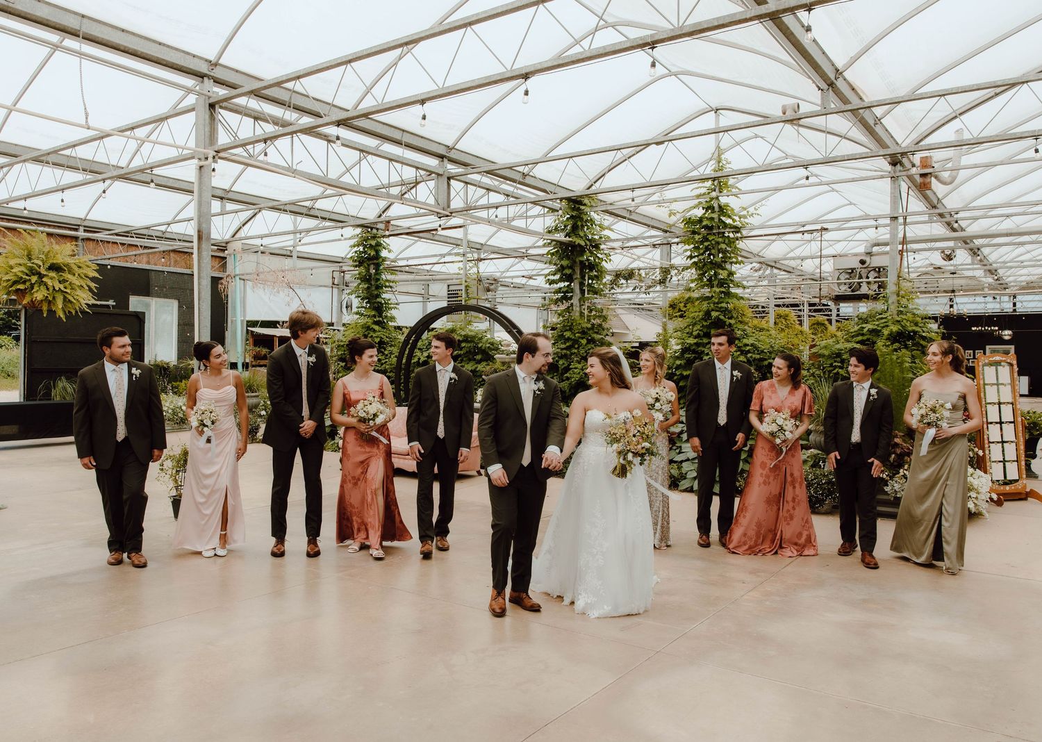 Wedding party walks together in a sunlit greenhouse venue, bride in white gown with bridesmaids in dusty rose and sage dresses.