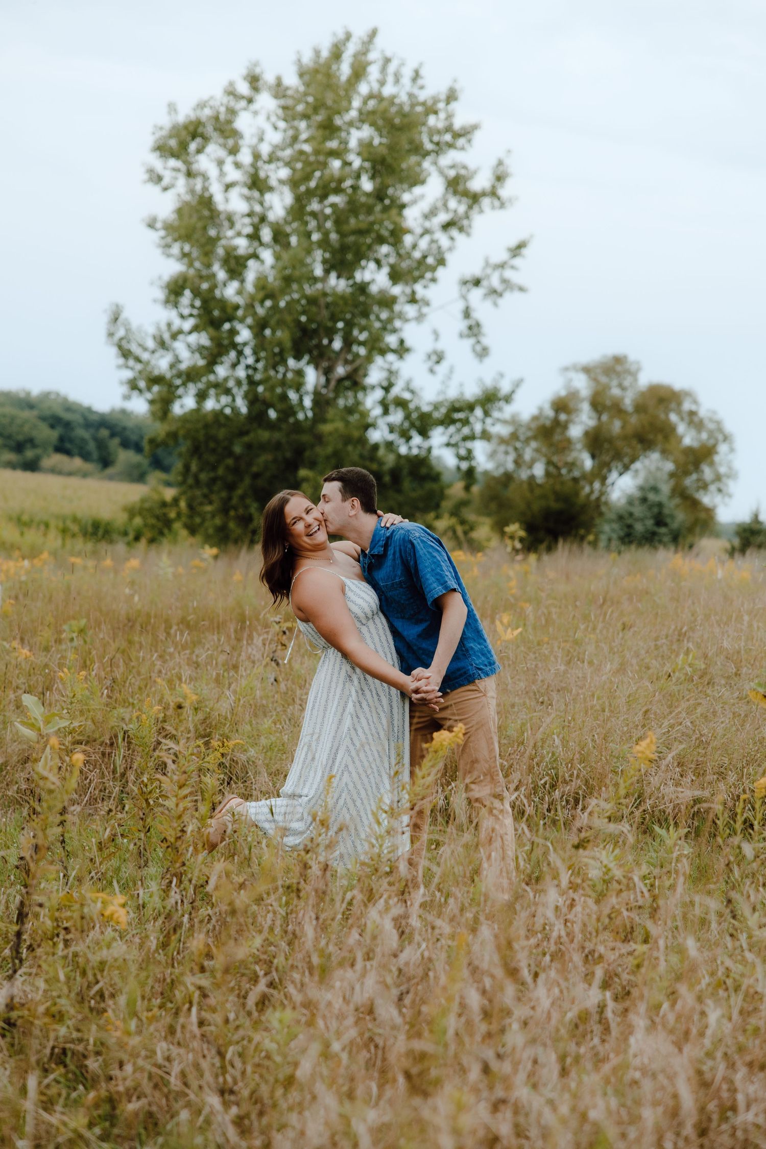 A couple shares a romantic kiss in a golden meadow, with lush green trees in the background on a cloudy day.
