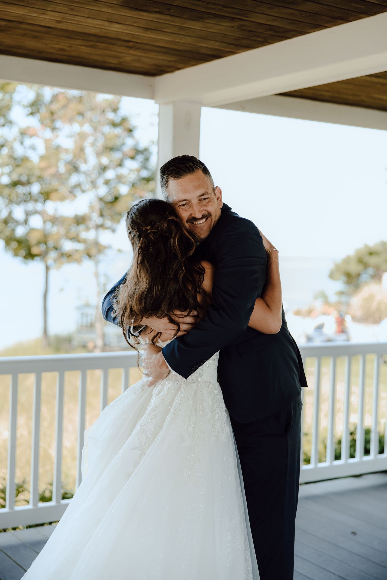 Groom in navy suit smiling while embracing bride in white lace gown on decorated porch.
