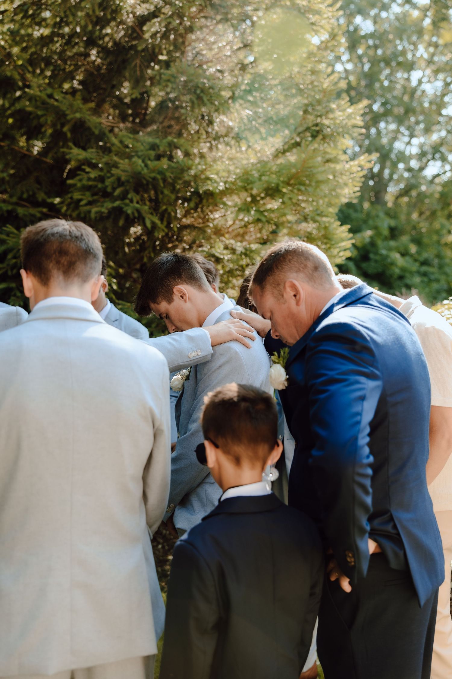 Groomsmen gather in prayer circle with groom in light blue suit before outdoor wedding ceremony.
