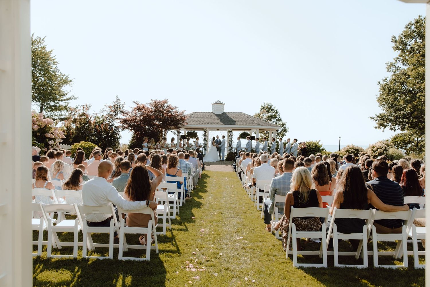 Outdoor wedding ceremony with guests seated in white chairs on a green lawn facing a gazebo altar under a clear blue sky.