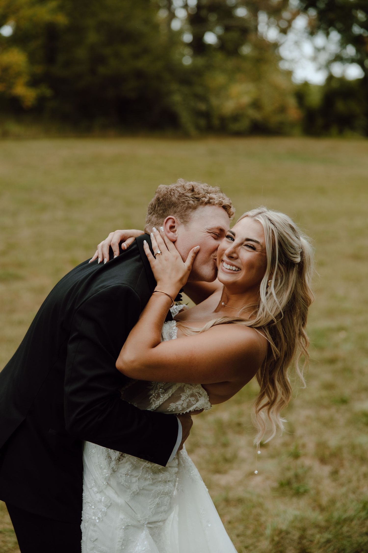 A groom in a black suit kisses his smiling bride in a white lace gown outdoors in a green field.