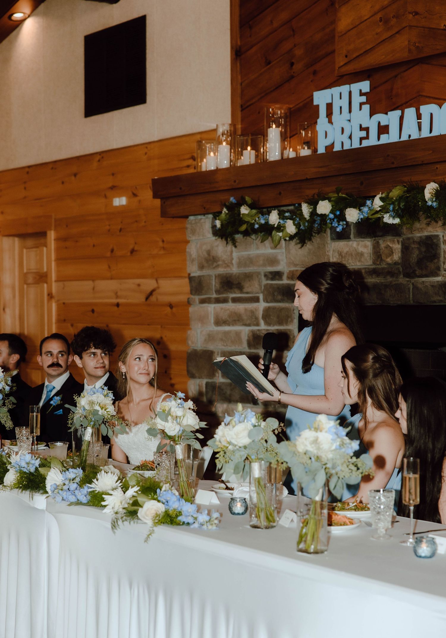 Wedding reception head table with guests listening to a bridesmaid speech near a stone fireplace.