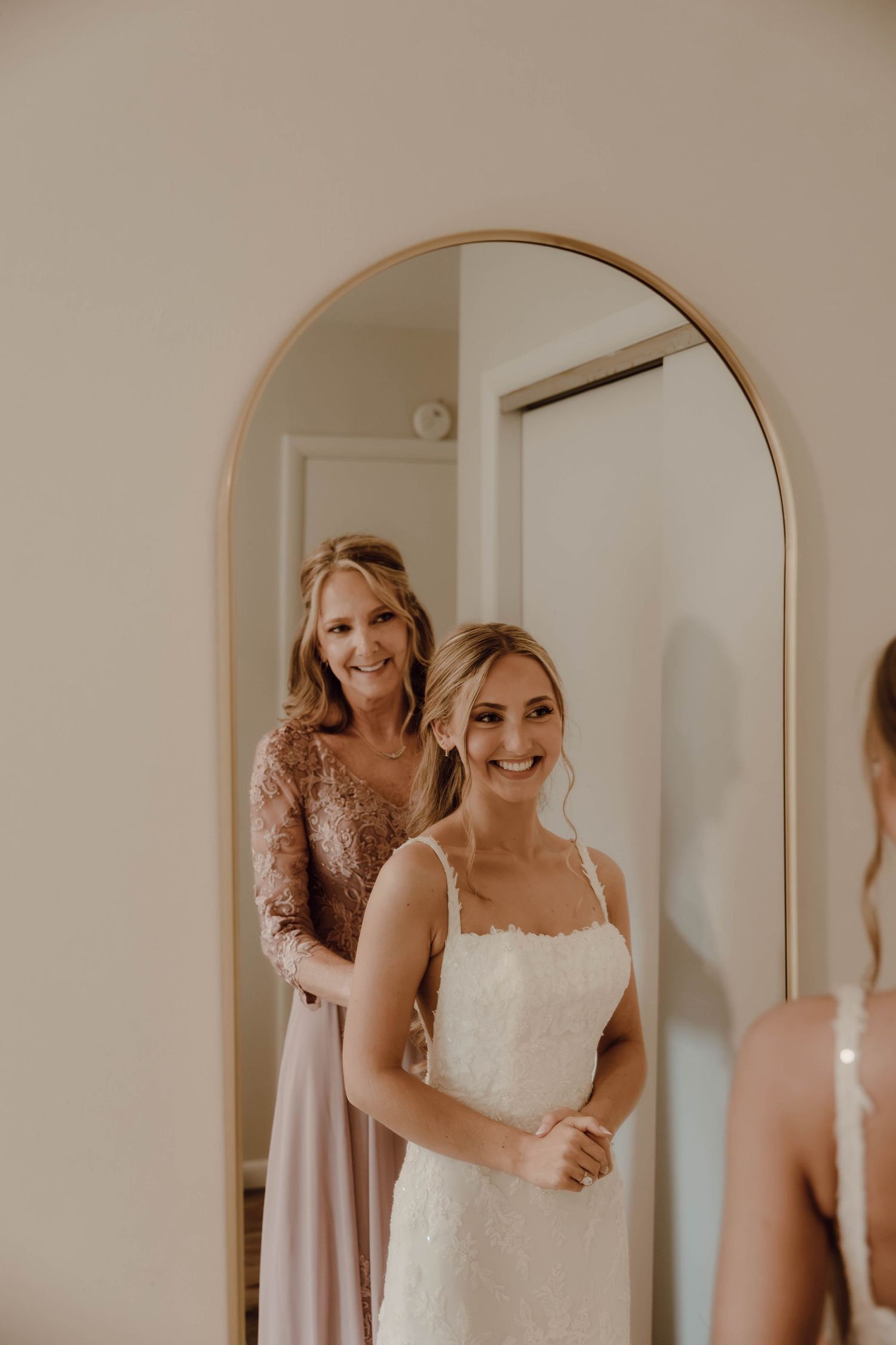 Bride smiling in mirror as bridesmaid in pink sequin gown helps with wedding dress fitting.