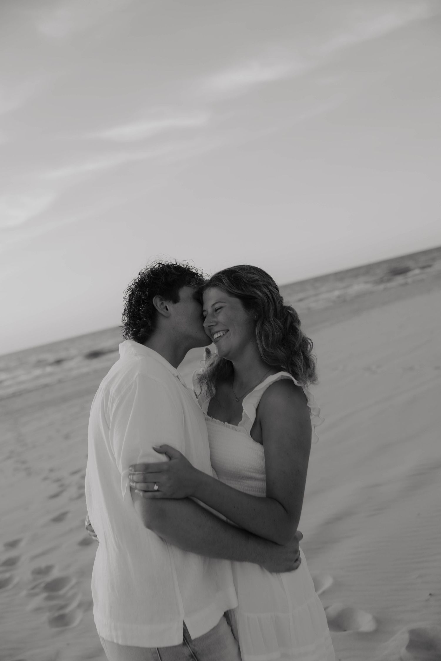 Black and white photo of a couple embracing and sharing a romantic moment on a sandy beach.