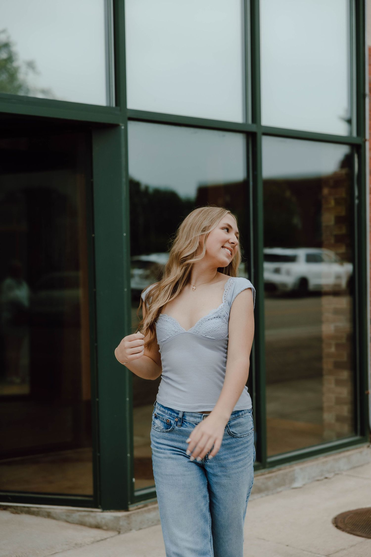 Young blonde woman in gray bodysuit and jeans smiling outdoors near large green-framed windows.