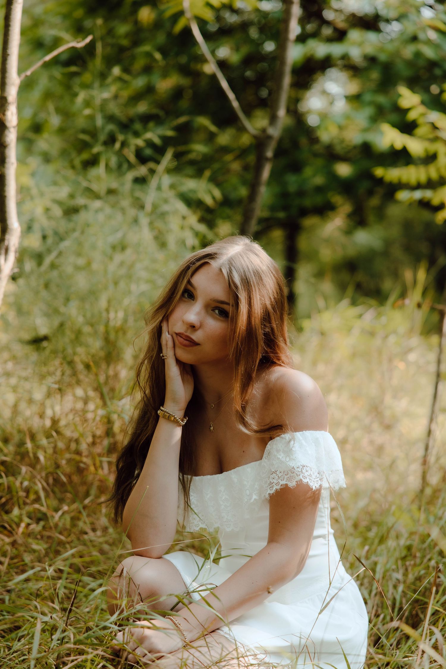 Young woman in white off-shoulder lace dress sitting gracefully in green forest grass among trees.