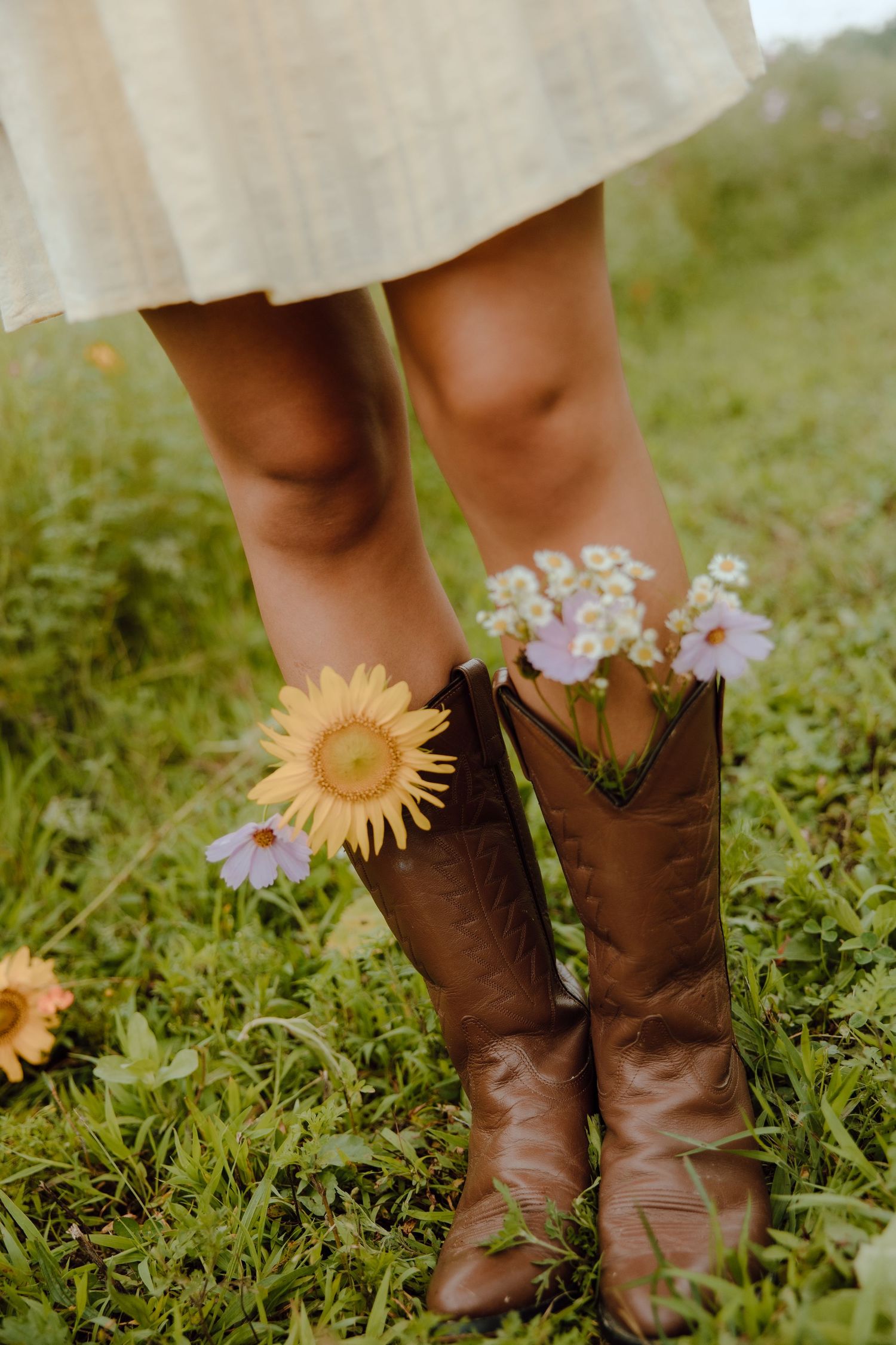 Person wearing brown cowboy boots filled with sunflowers and wildflowers, standing on green grass in a cream dress.