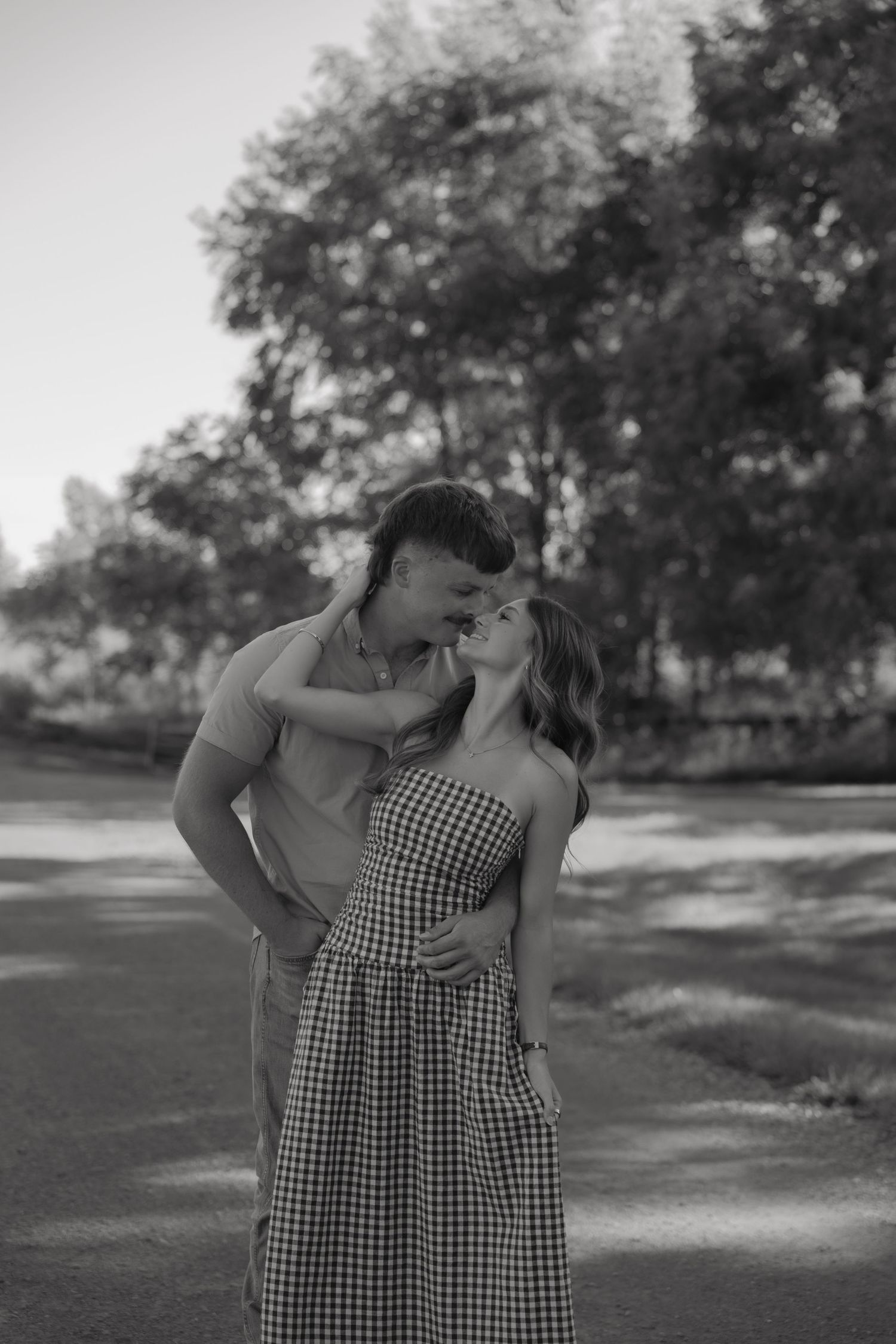 Black and white photo of a romantic couple embracing and almost kissing in a sunny park setting.
