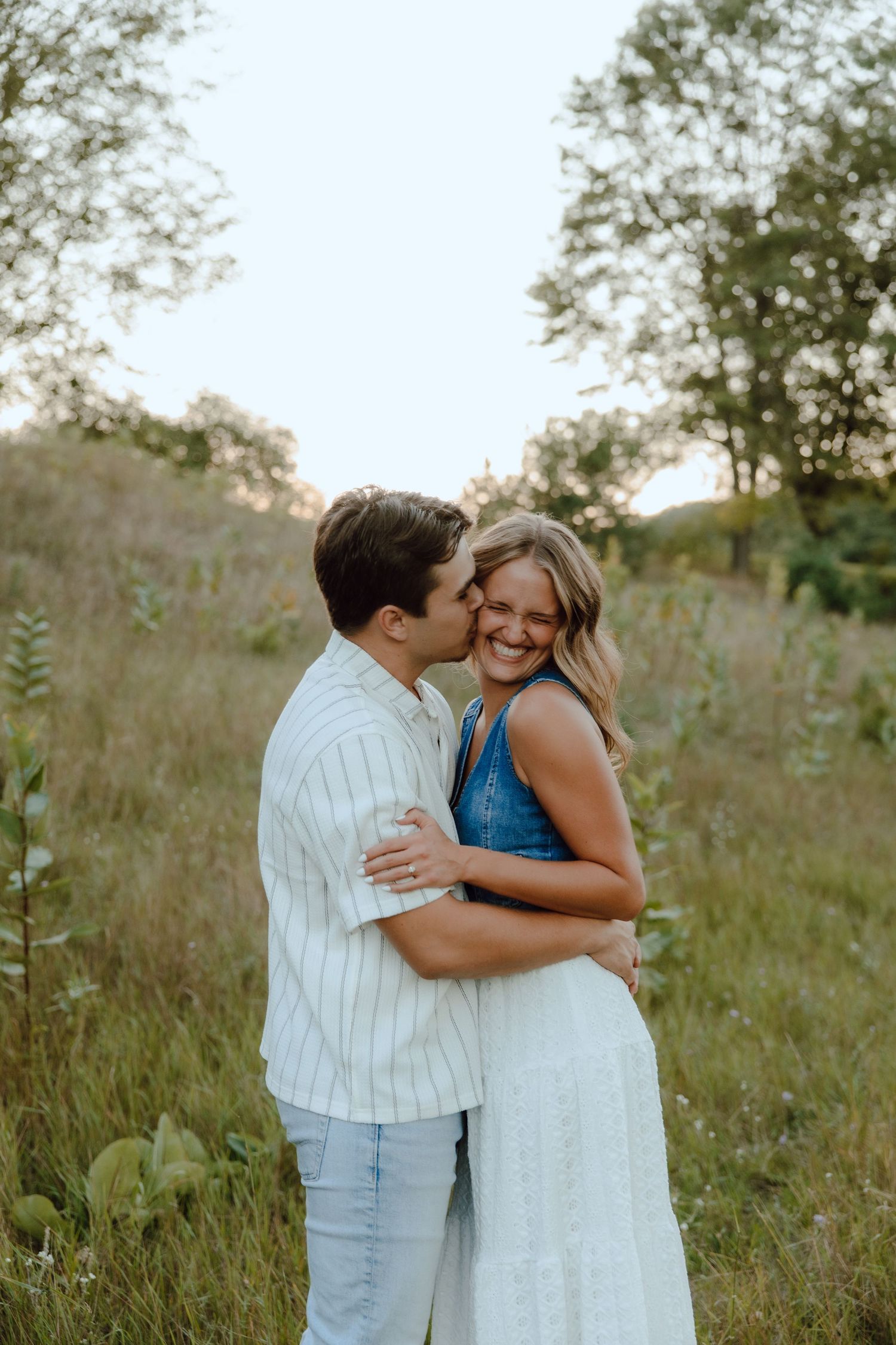 A couple embraces in a lush green meadow, the man kissing the woman's cheek as she smiles joyfully.