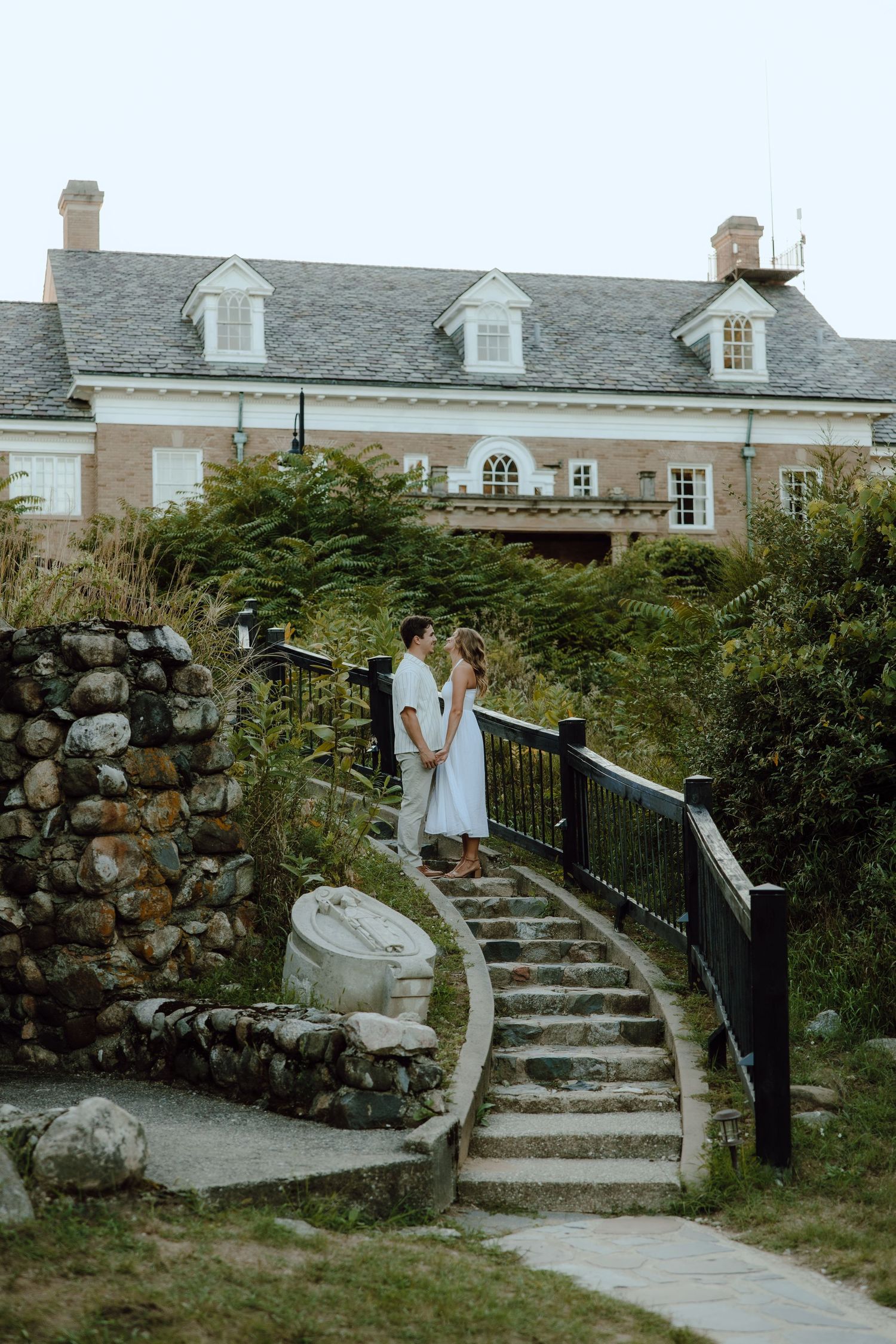 Couple embracing on stone steps beside a historic brick manor surrounded by lush greenery.