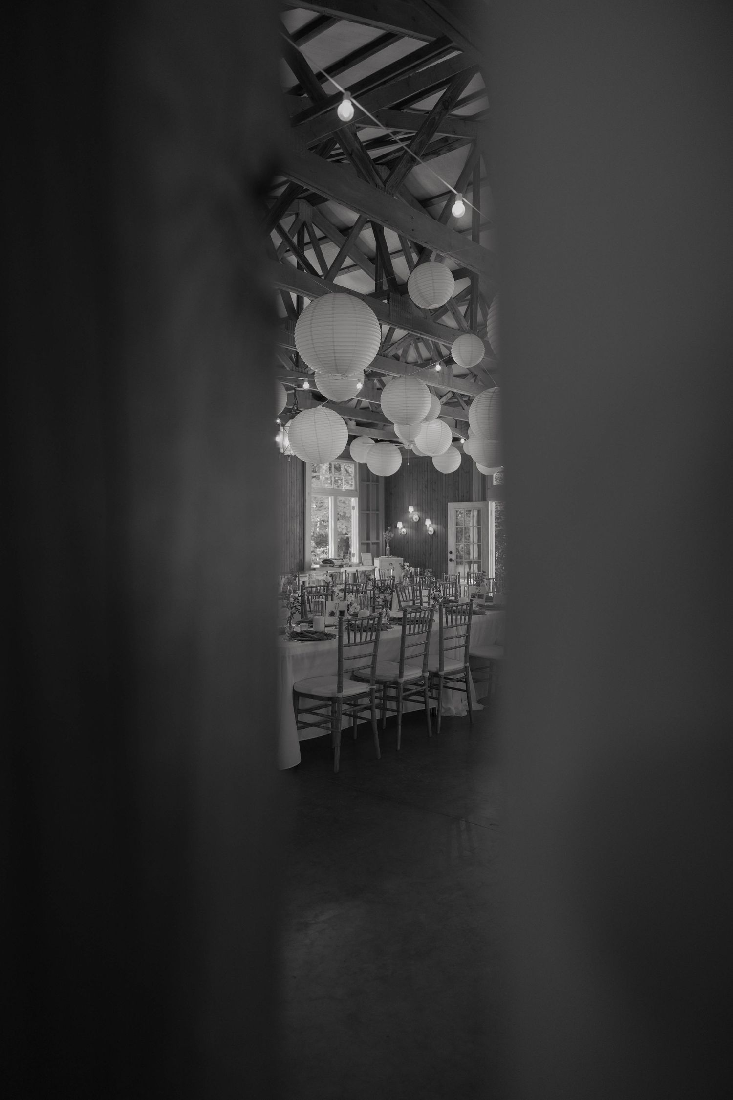 Black and white photo of elegant banquet hall with round paper lanterns hanging from wooden beamed ceiling over long dining tables.