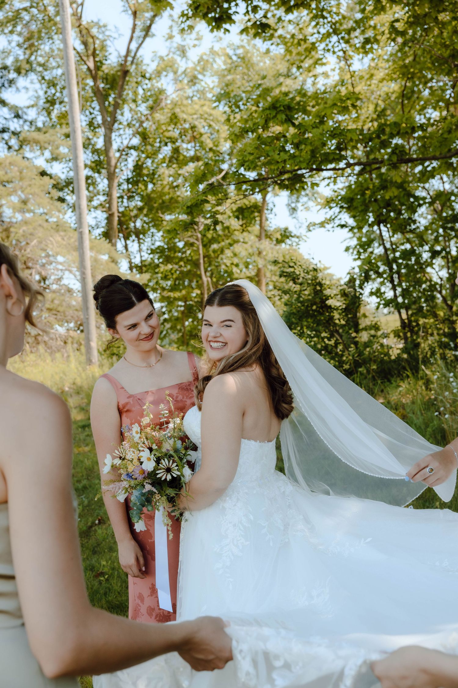 Bride in white lace gown and veil smiling with bridesmaid in pink dress outdoors among green trees.