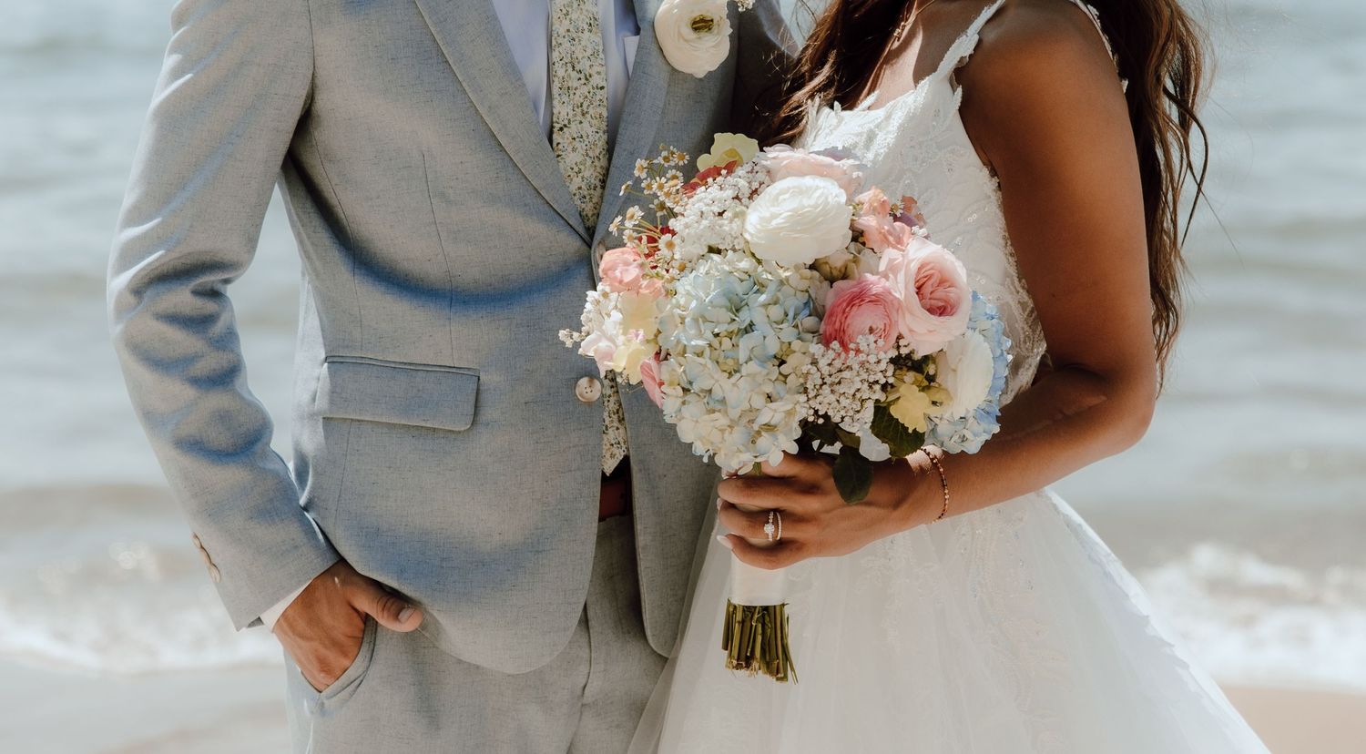 Bride and groom standing together at beach wedding, holding pastel pink and blue flower bouquet.