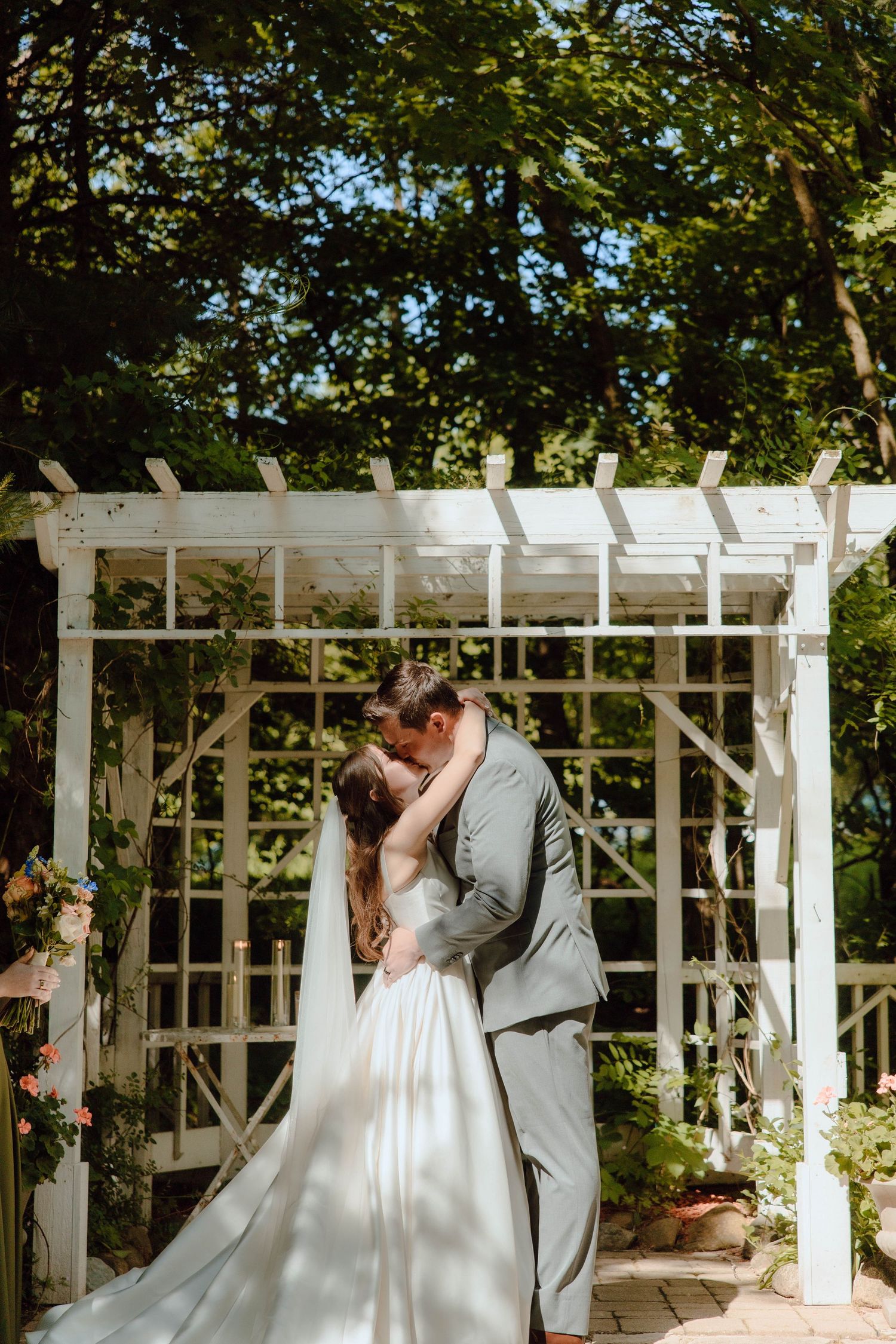 Bride and groom share their first kiss under a white garden pergola surrounded by lush greenery.
