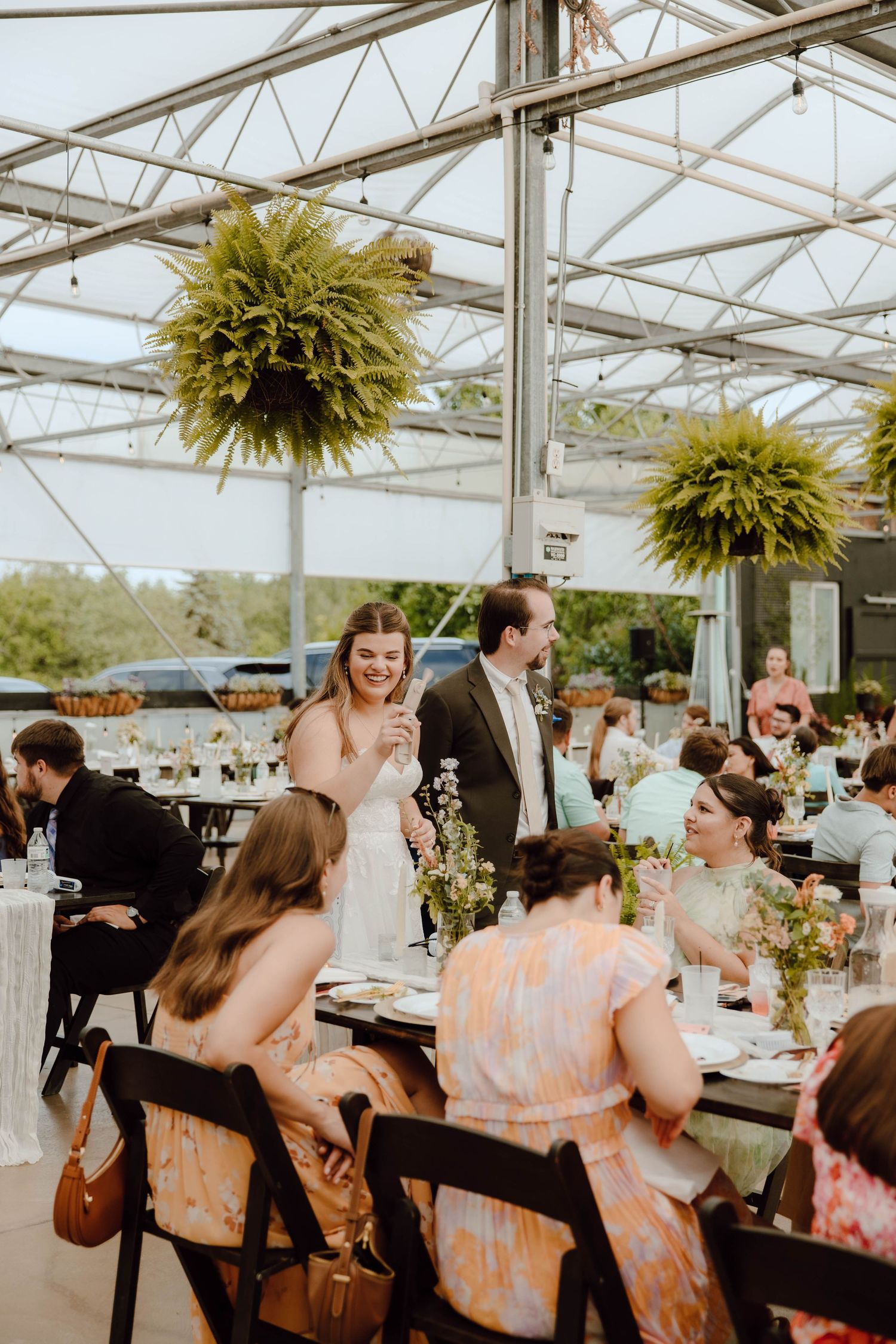 Bride and groom mingle with guests during elegant greenhouse wedding reception with hanging ferns.