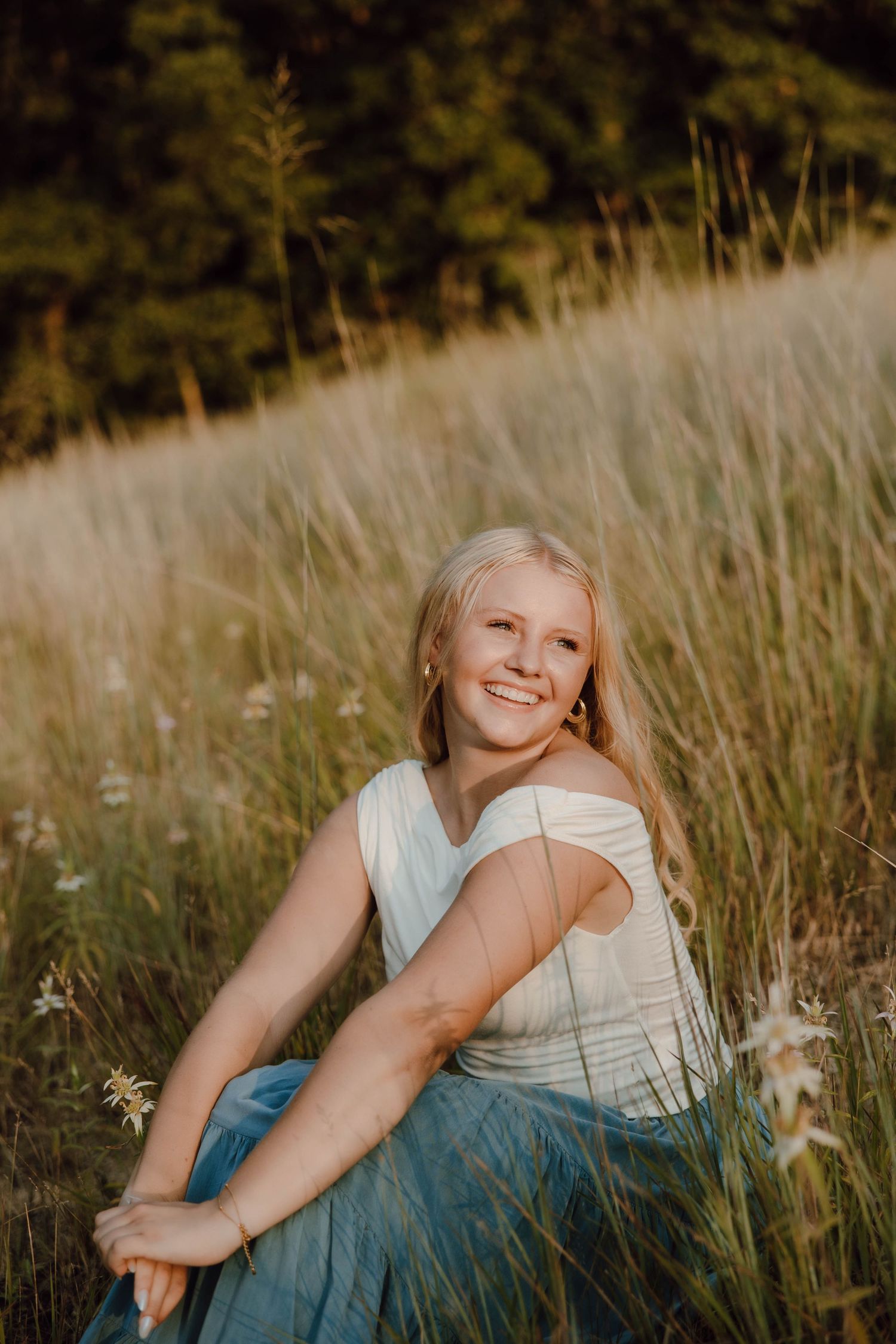 Smiling blonde teen girl in white top and blue skirt sitting in golden meadow grass with wildflowers.