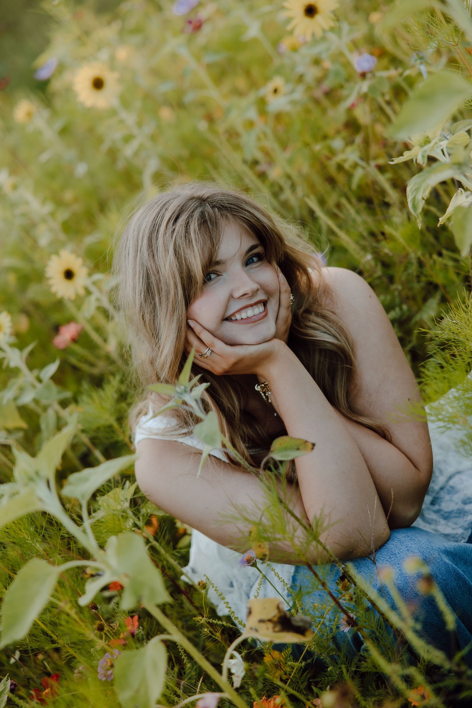 Smiling blonde woman lying in a wildflower field with sunflowers, resting her chin on her hand.