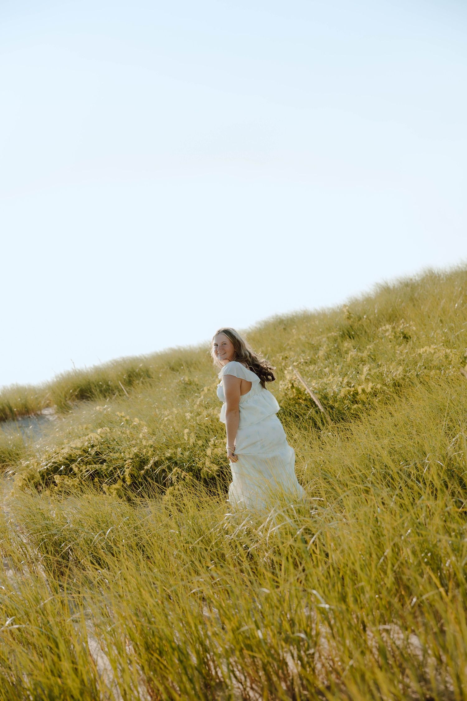 Woman in white dress standing on grassy sand dune with wind blowing her hair against a clear sky.