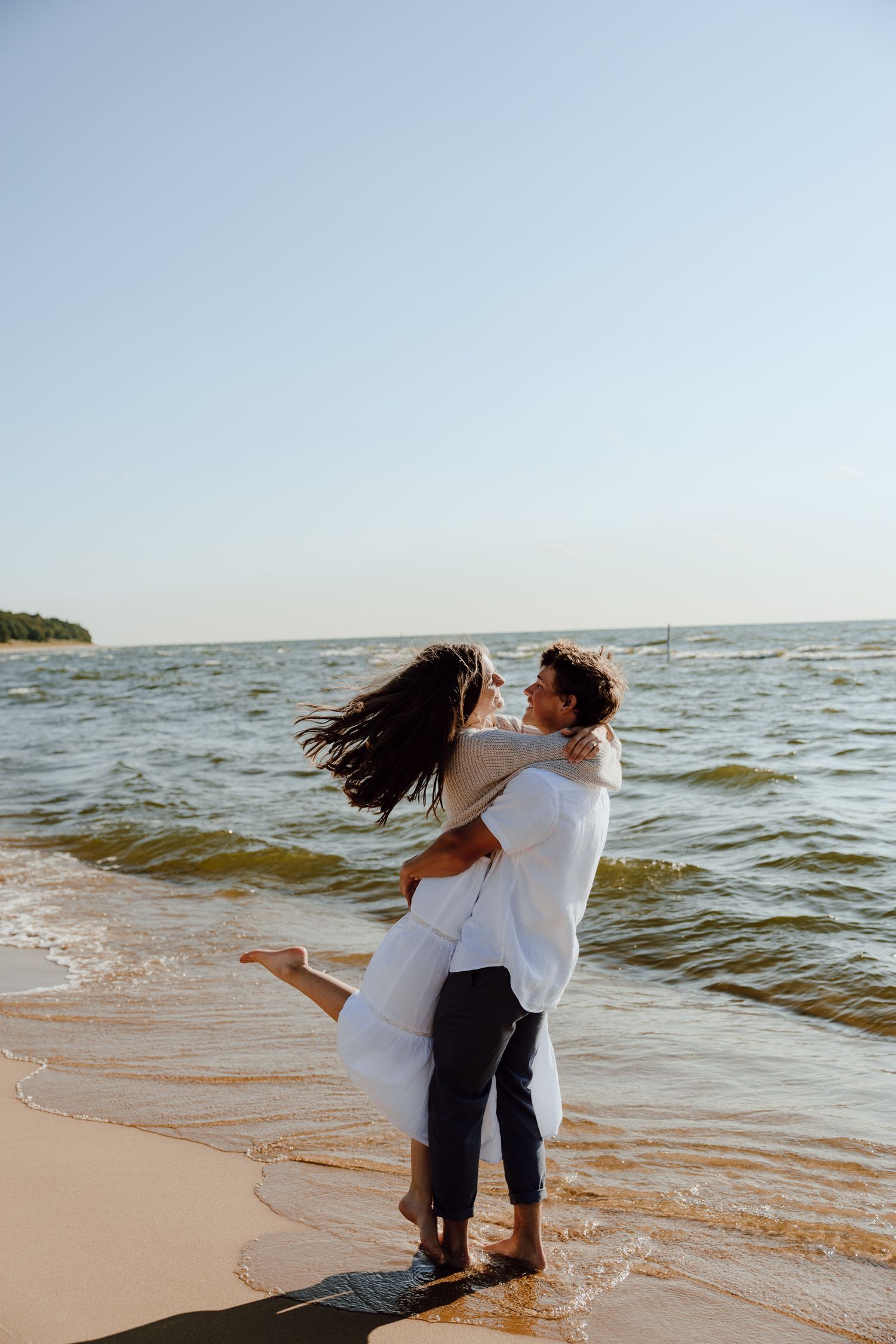 Playful couple embraces on a sandy beach with waves crashing behind them.
