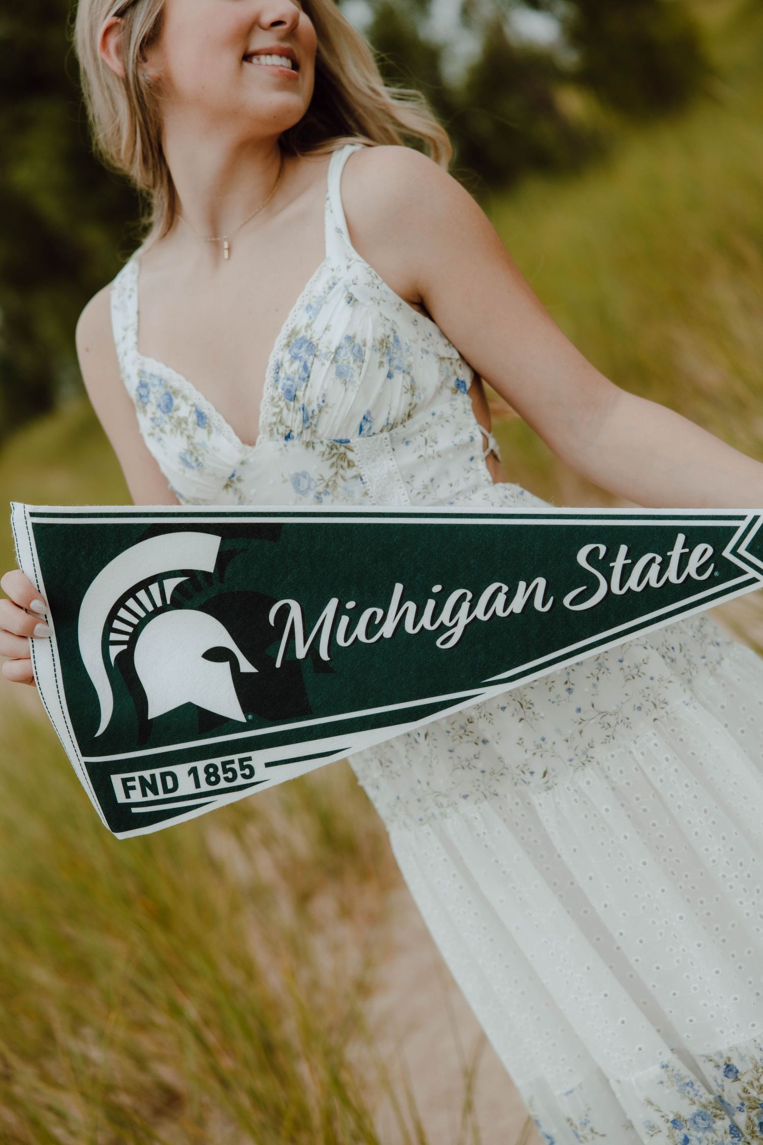 Smiling blonde woman in floral dress holding a green Michigan State University pennant flag outdoors.
