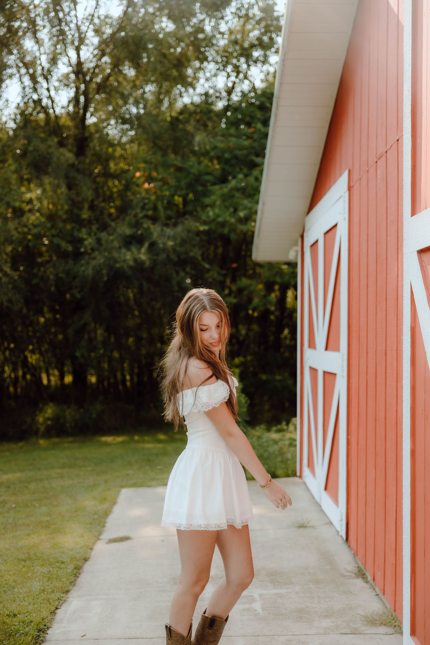 Young woman in white off-shoulder mini dress and brown cowboy boots posing beside a red barn outdoors.