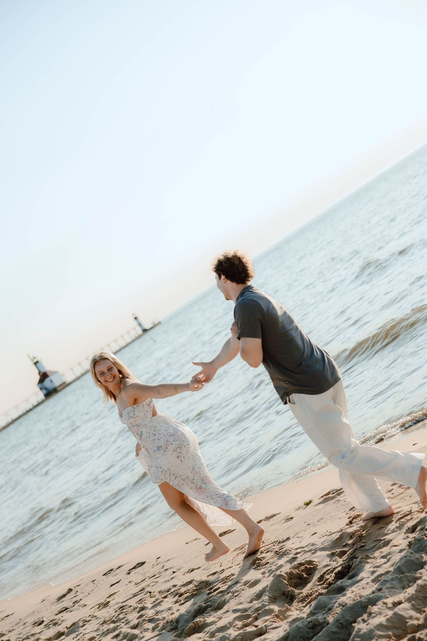 Couple holding hands and dancing playfully on a sandy beach at sunset near the water.