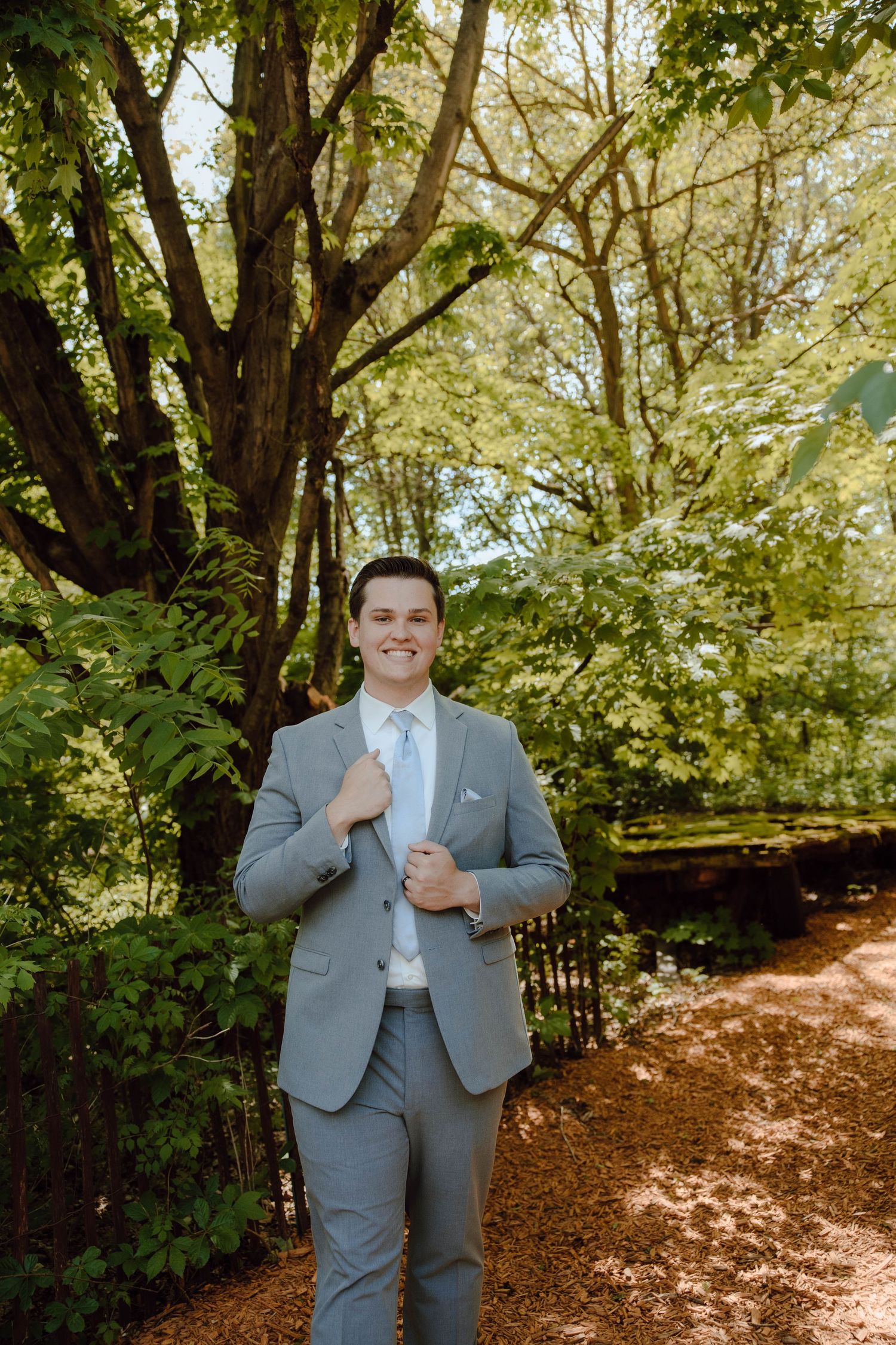 Handsome groom in a light gray suit smiles confidently while standing in a lush green garden.