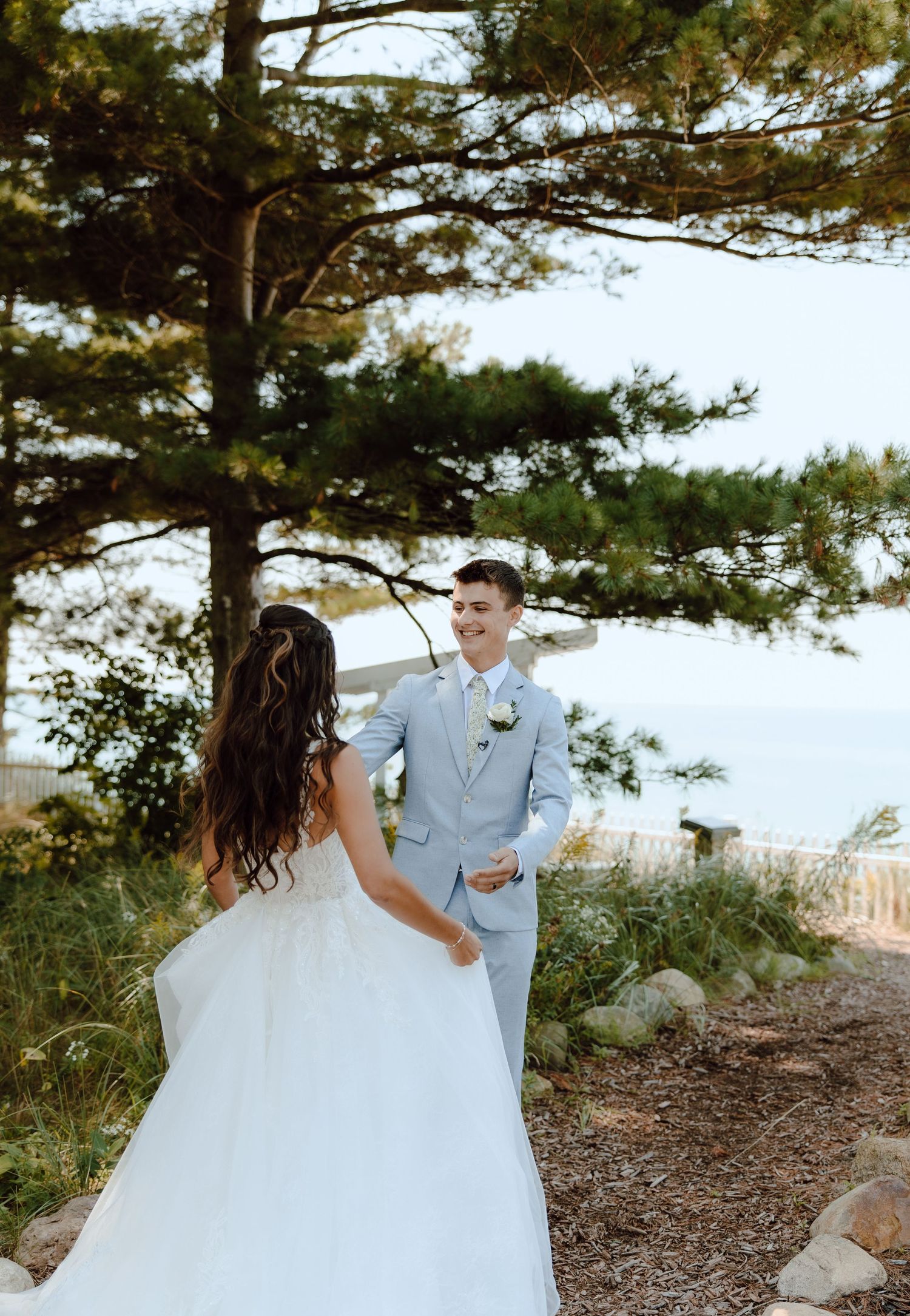 Groom in light blue suit smiling at bride in white gown during outdoor first look by pine trees.