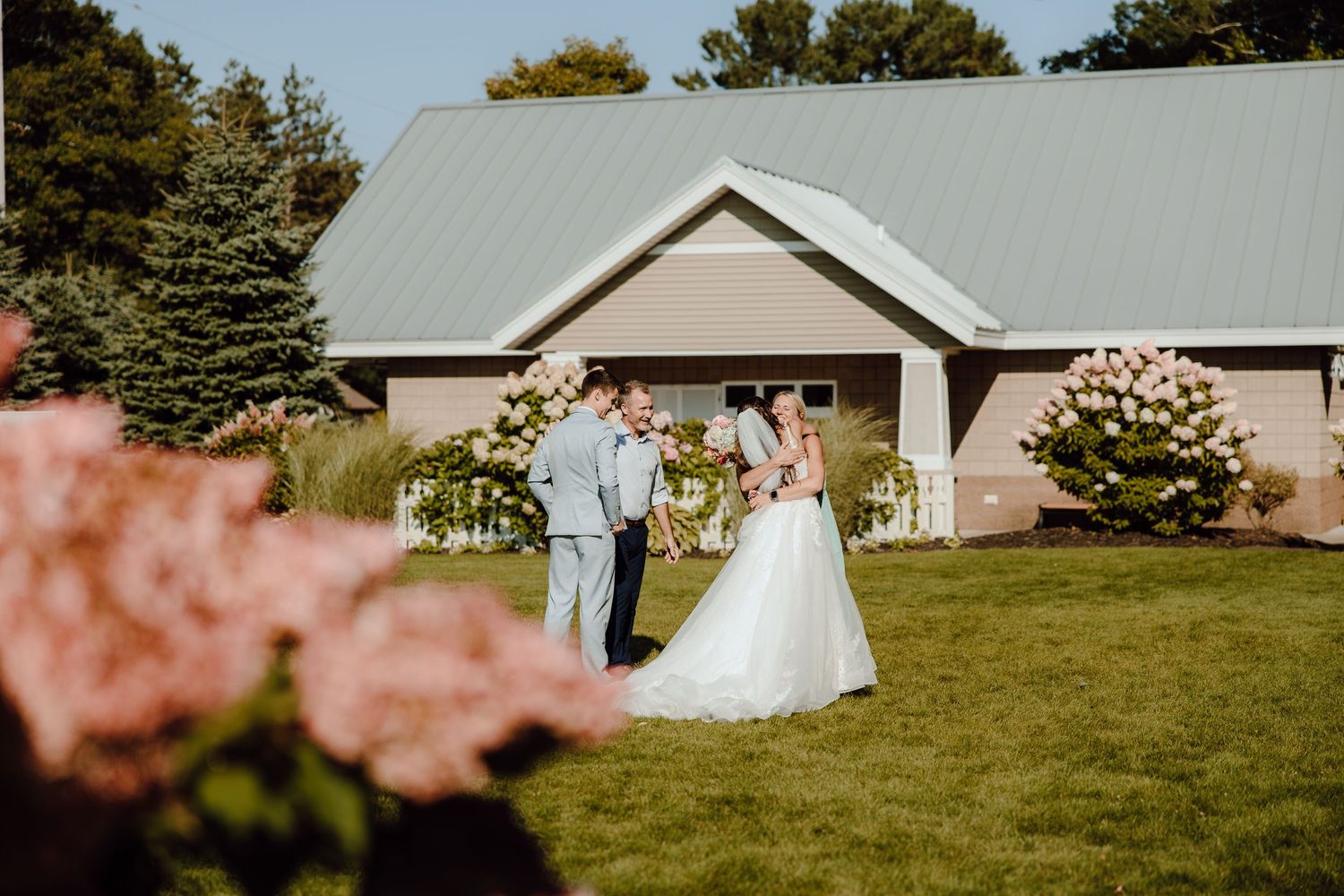 Bride and groom share intimate moment on green lawn beside rustic venue with pink hydrangeas.
