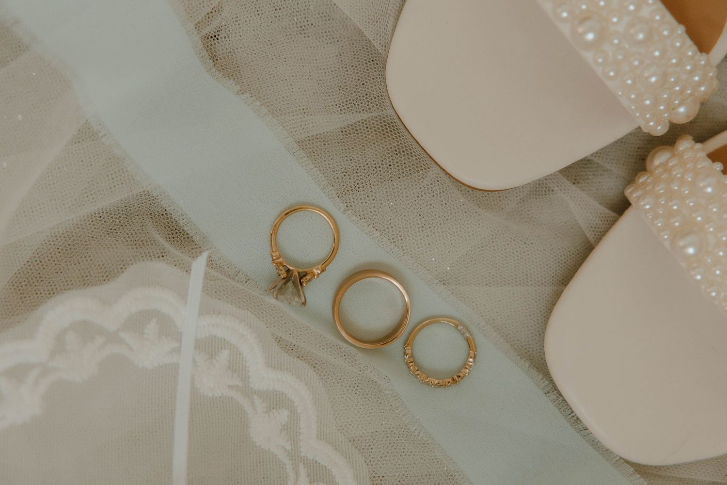 Gold wedding rings and bands displayed on a white ribbon beside pearl-embellished bridal heels on decorative tile.