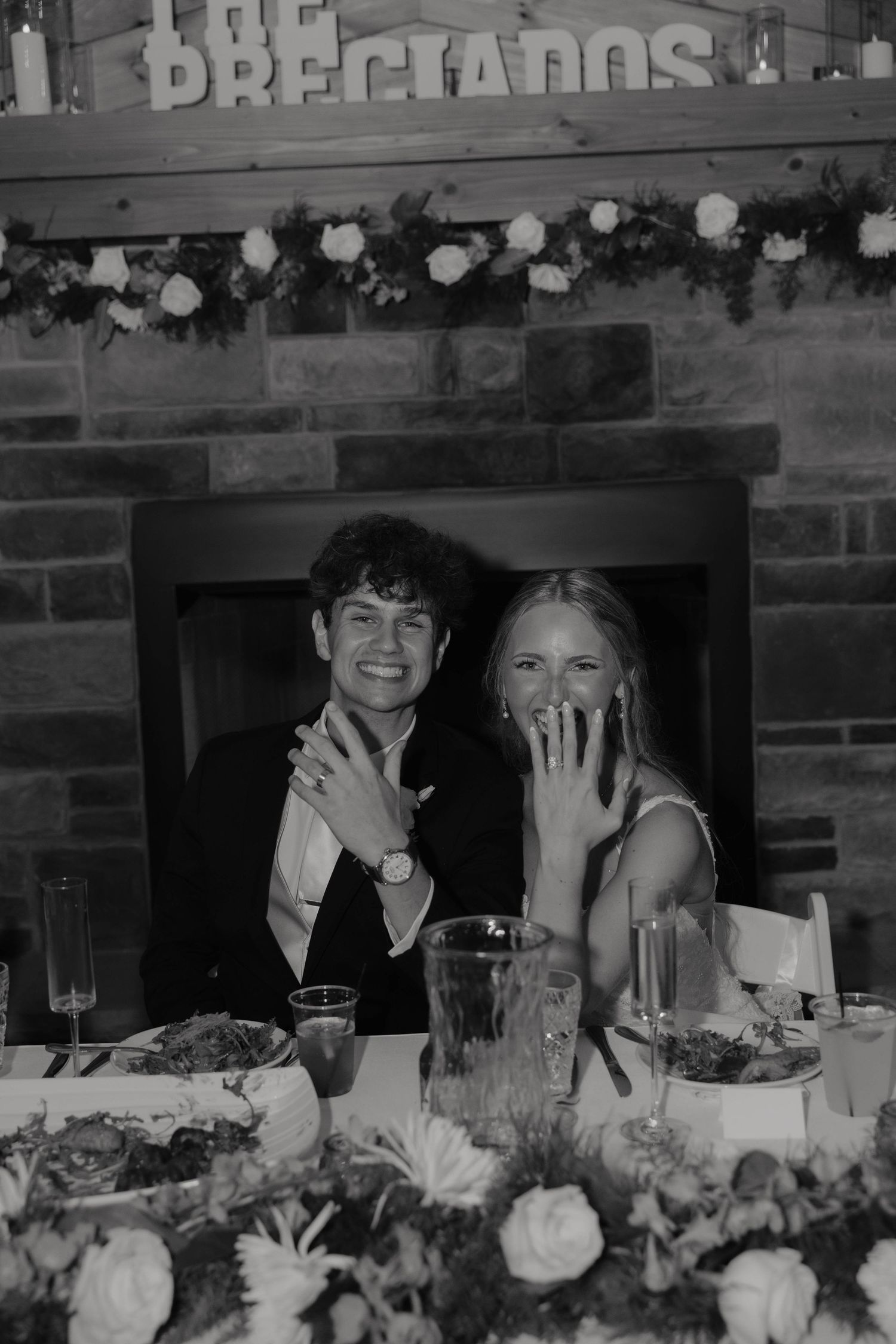 Newlyweds showing off wedding rings at reception table beneath 