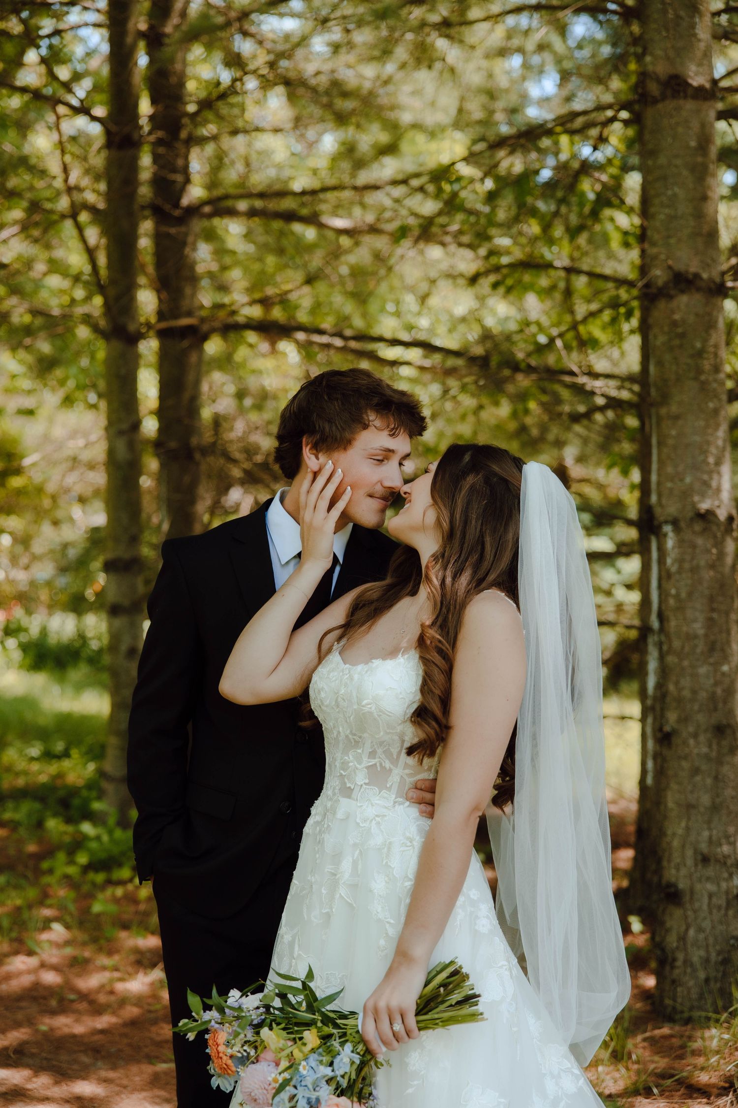 A bride and groom share a kiss in a sunlit forest setting.