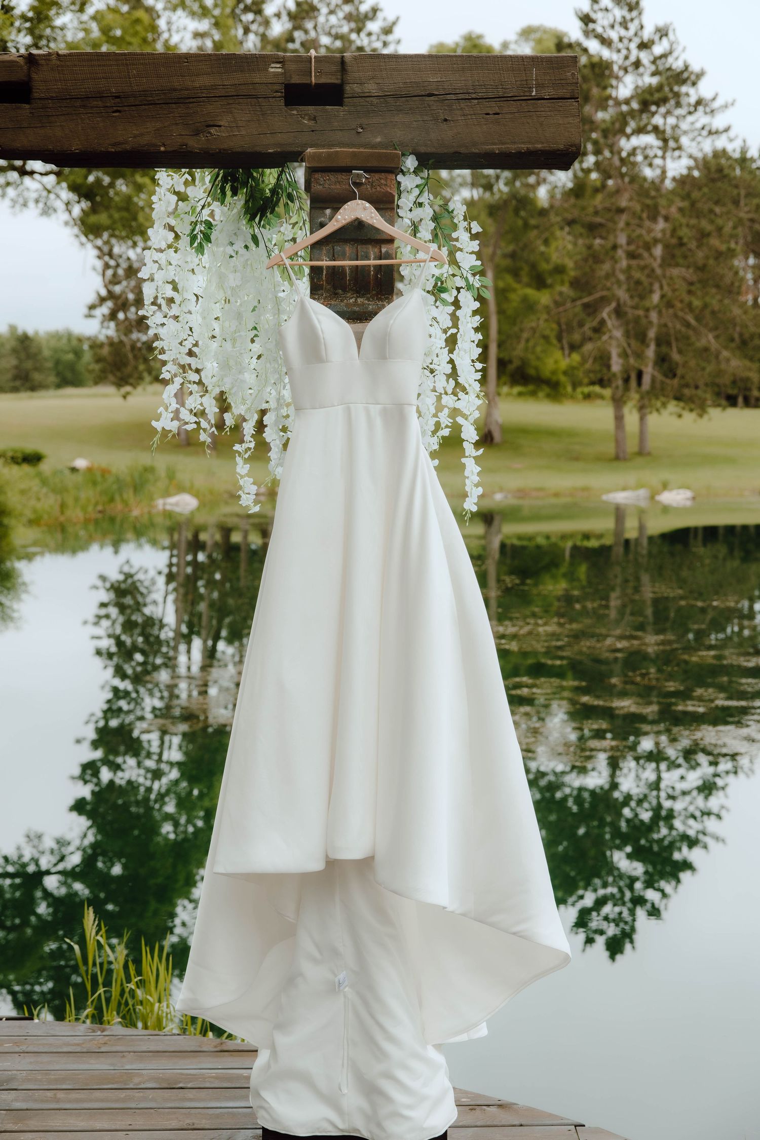 Elegant white wedding dress hanging on a wooden pergola over a serene reflective pond outdoors.