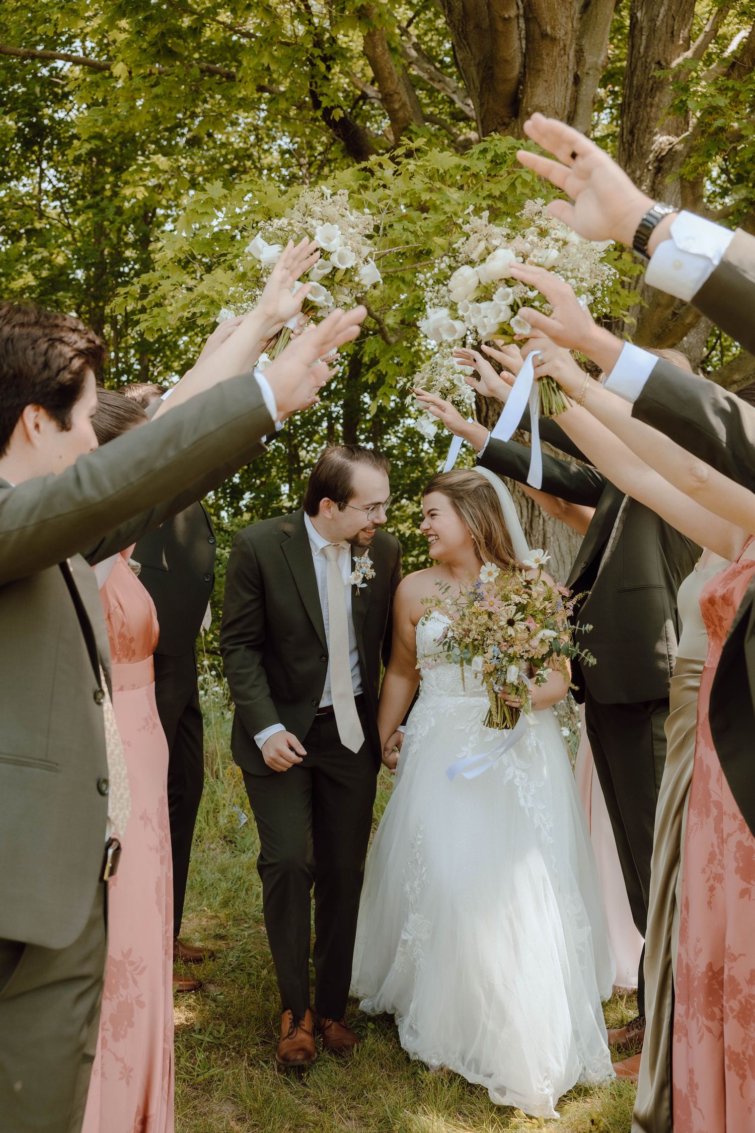 Bride and groom walk through an arch of raised hands and flowers held by wedding party outdoors.