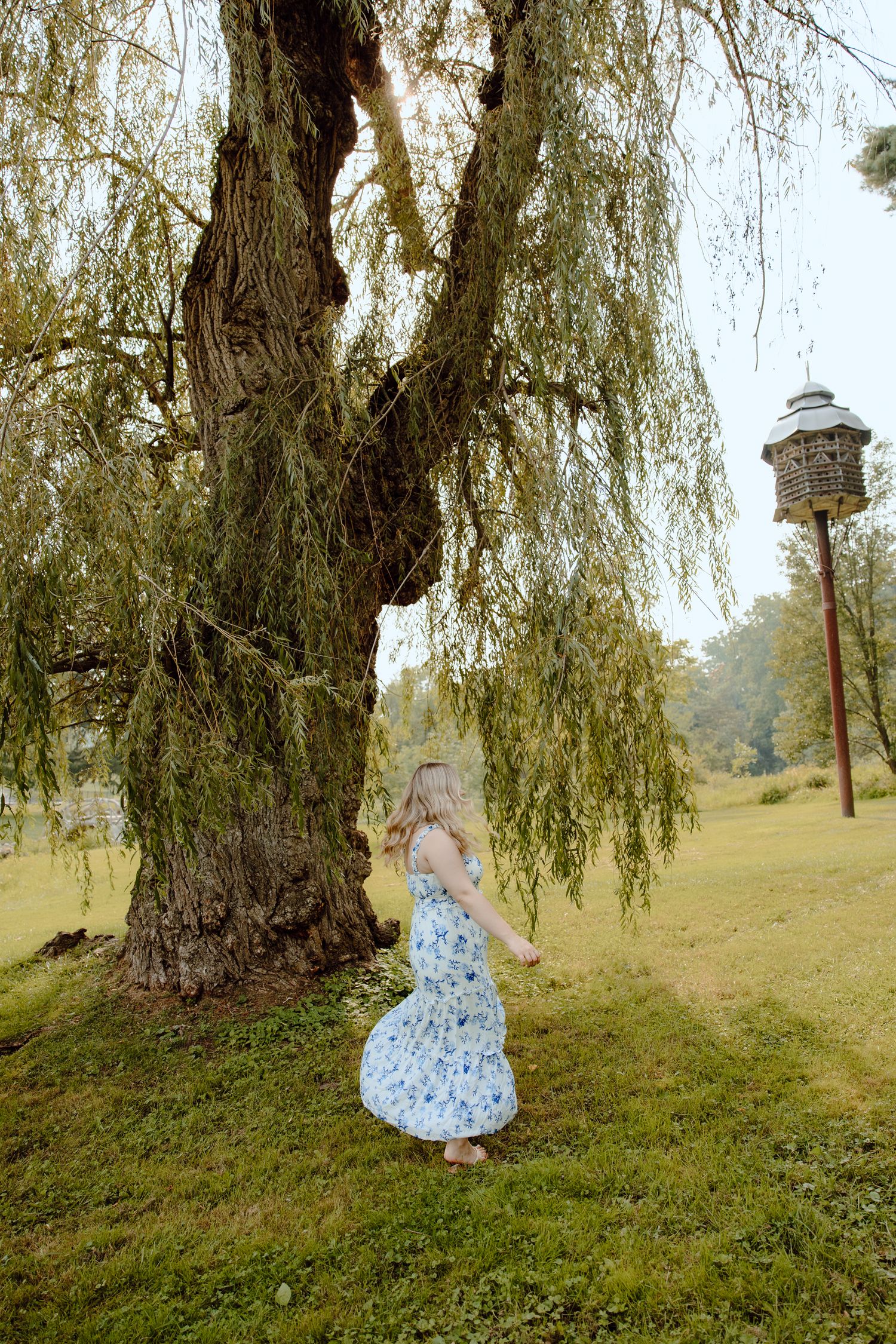 Person in floral dress poses whimsically around a large willow tree.