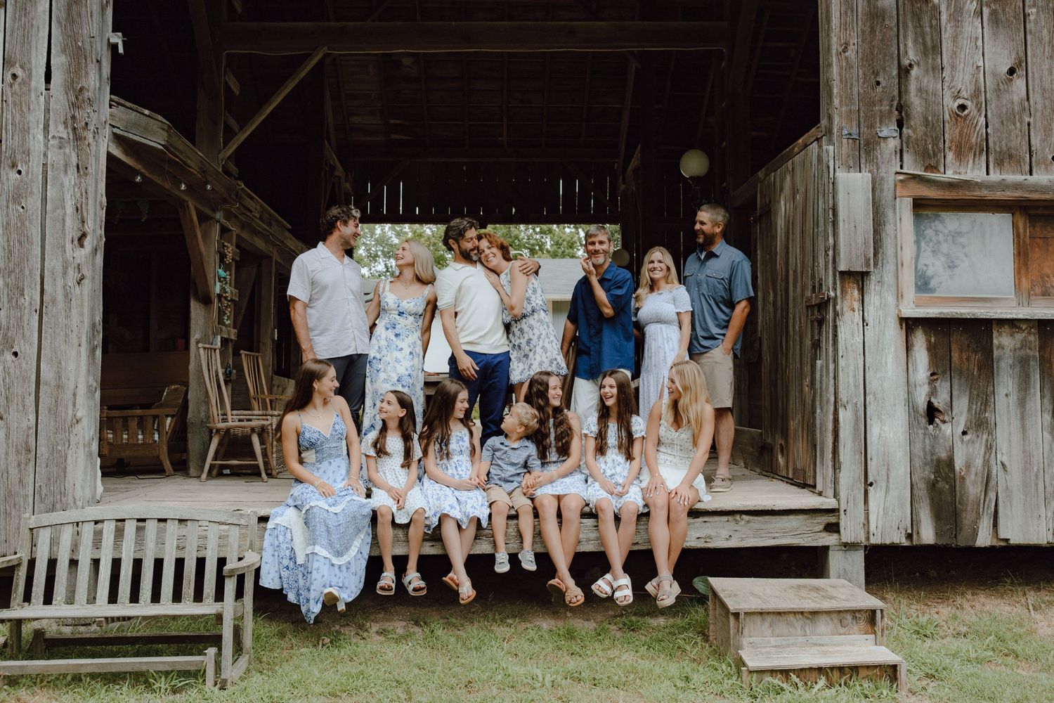 Extended family gathering posed casually outside rustic barn wearing matching blue and white attire.