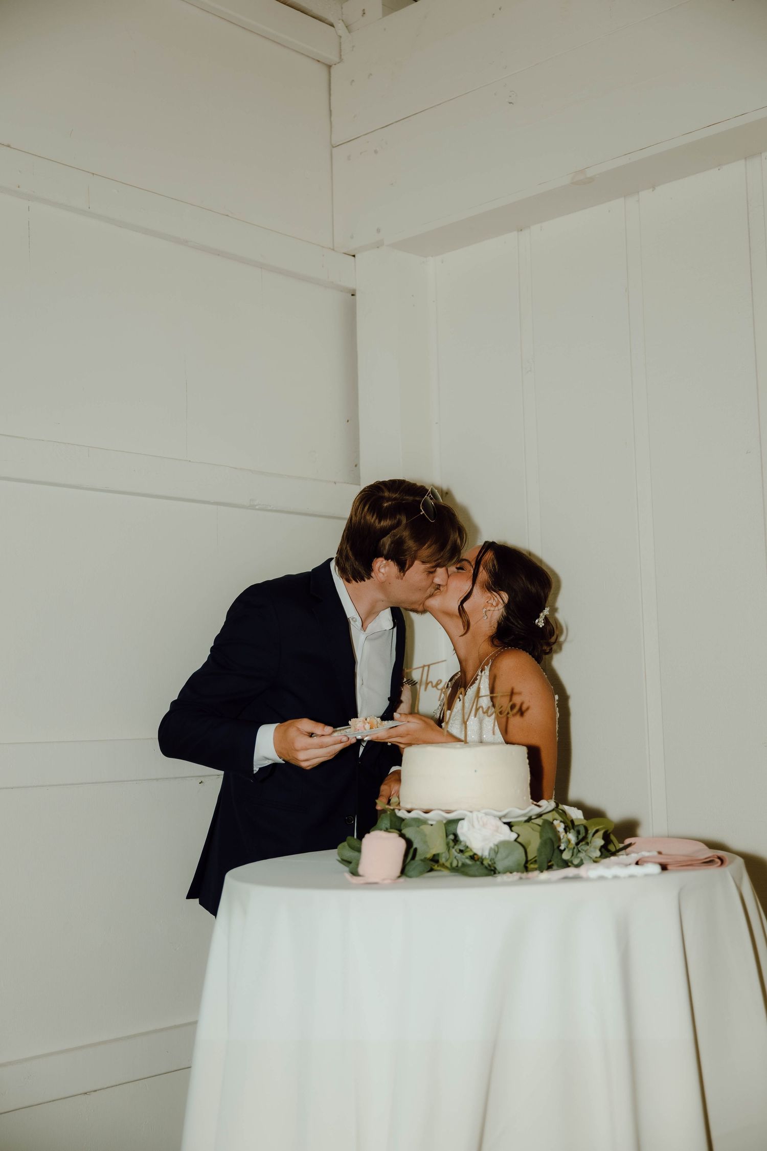 Bride and groom sharing a kiss while cutting their two-tier white wedding cake at reception.