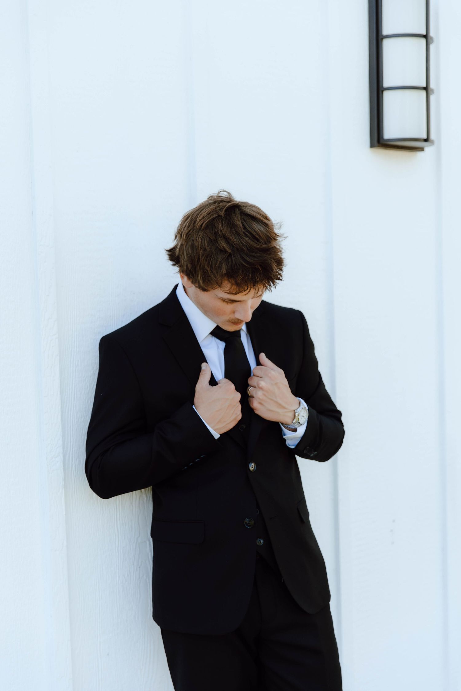 A groom in a black suit adjusts his tie against a white wall.