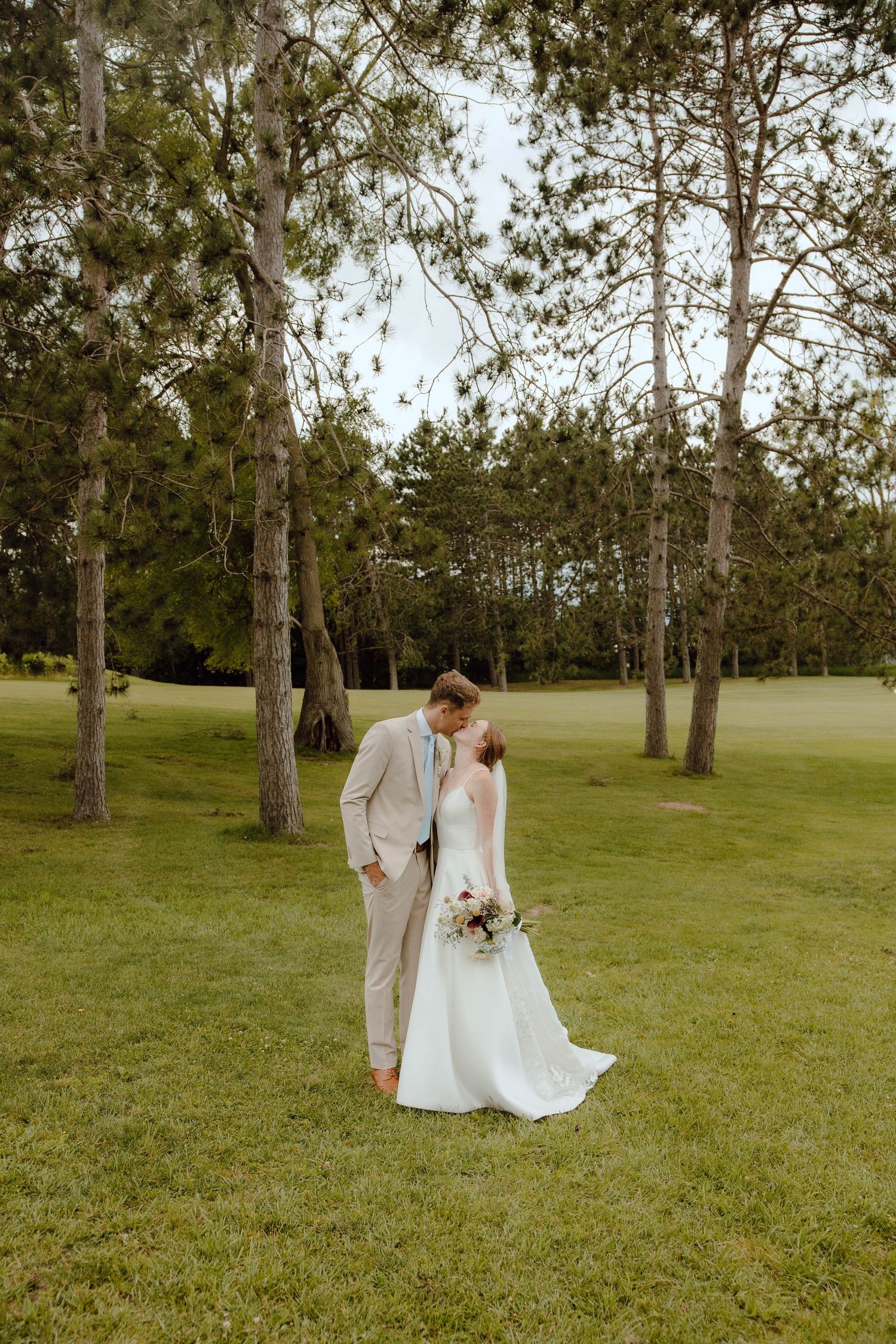 Bride and groom share a romantic kiss outdoors on lush green grass surrounded by tall pine trees on their wedding day.