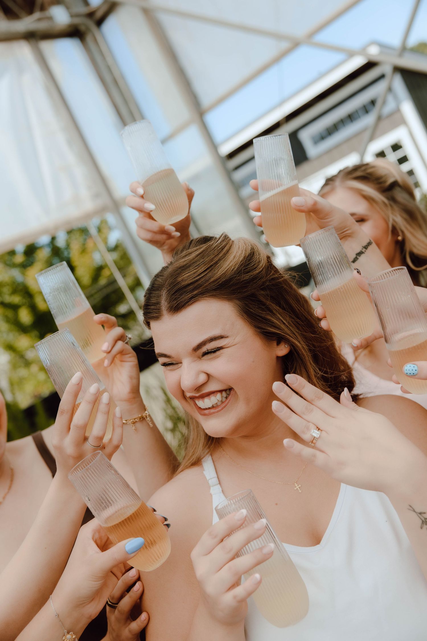Joyful bride laughing as bridesmaids toast with champagne glasses outdoors.