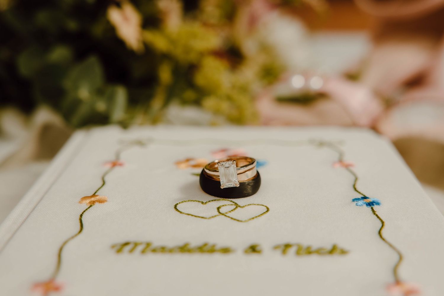 Wedding rings resting on a personalized white book embroidered with two names and colorful heart details.